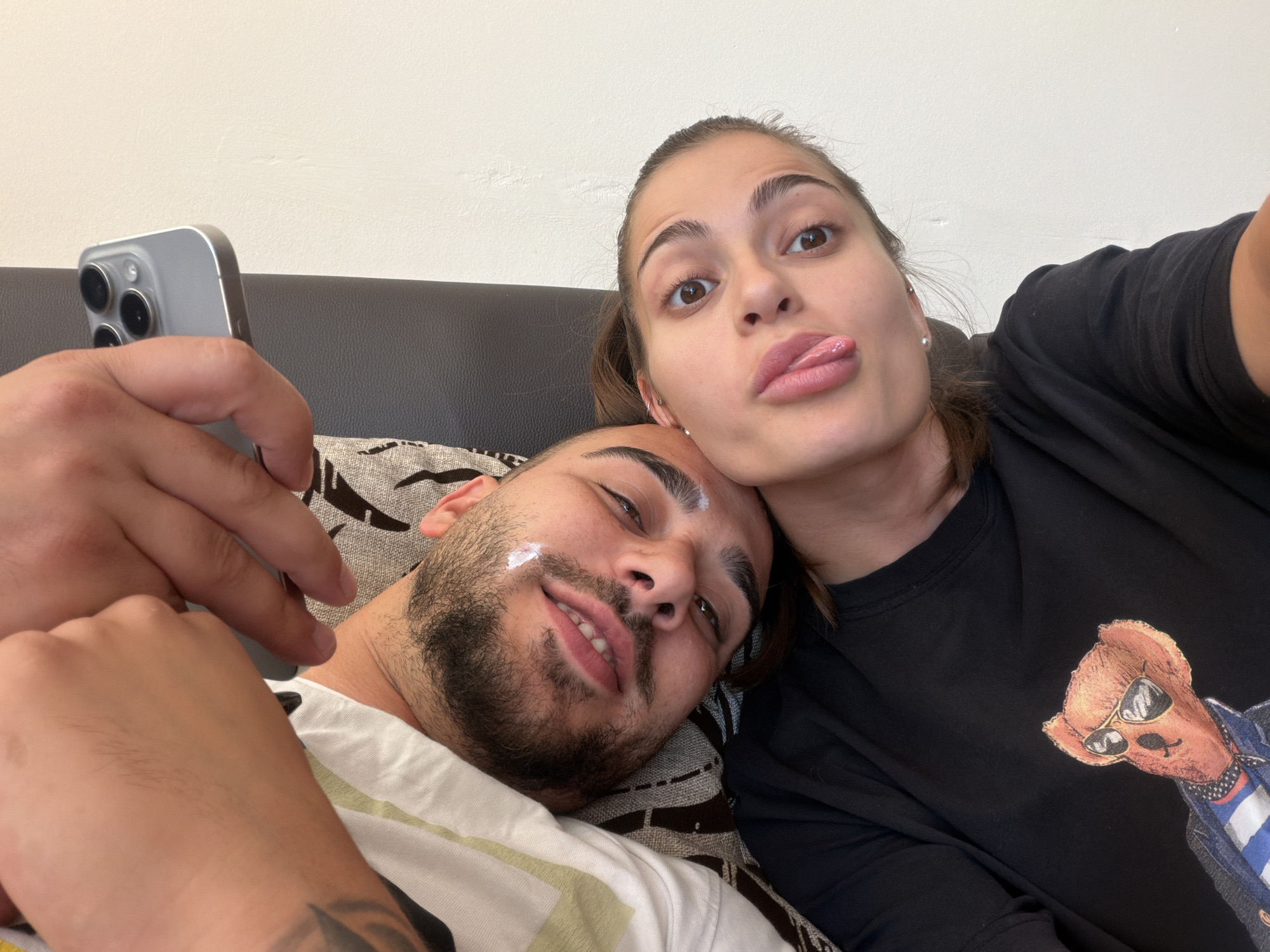 A close-up, selfie-style photograph captures two young adults lounging together, seemingly on a couch. The person in the foreground, on the left, is a man with dark hair, a beard, and light-colored smudges on his face. He is wearing a white t-shirt with green and yellow stripes and appears to be smiling. His right arm is visible, holding a silver smartphone. The woman on the right, with her hair pulled back and wearing a black t-shirt featuring a graphic of a teddy bear wearing sunglasses and a denim jacket, is sticking her tongue out at the camera. The background consists of a plain wall. The overall impression is one of playful intimacy.