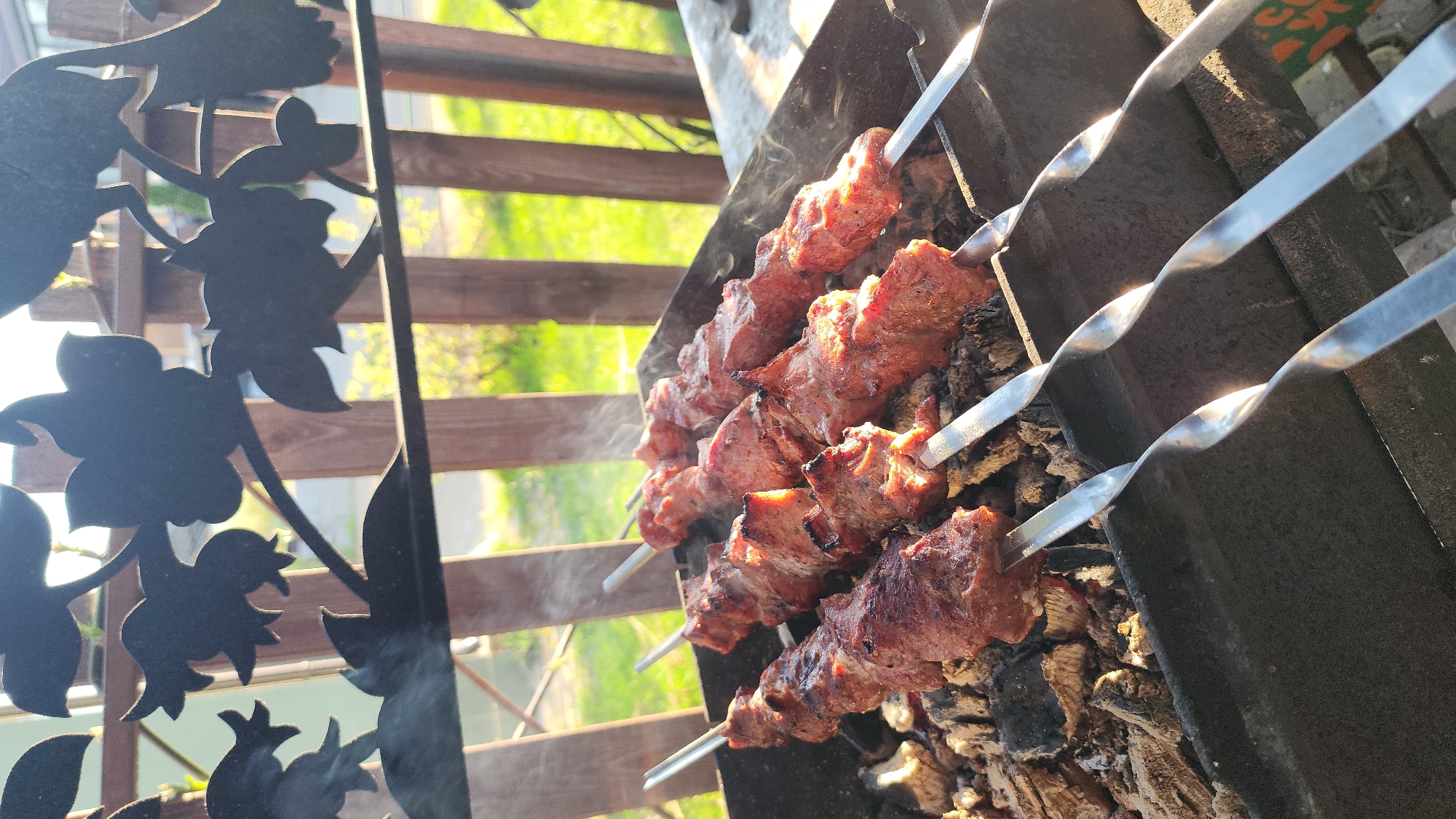 The image depicts a barbecue scene in Рыбхоз, Russia. Several metal skewers, each loaded with chunks of cooked meat, are laid across a charcoal grill. Smoke rises from the meat. In the background, a wooden fence and foliage are visible. A decorative metal screen with a floral design is in the foreground.
