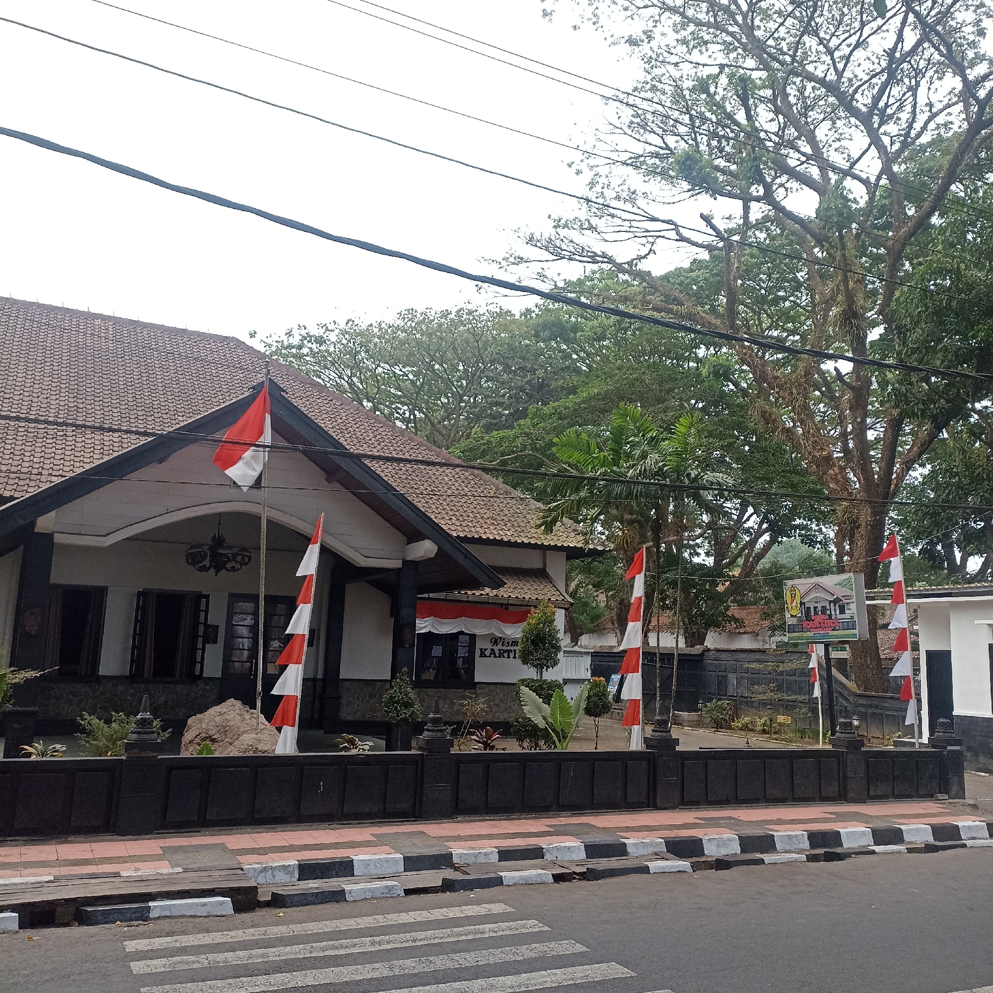 The image shows the front of a building with a tiled roof and a dark-colored facade. Indonesian flags, red and white, are displayed on flagpoles in front of the building. The building appears to be a historical site or museum, possibly "Wisma Kartini" as indicated by signage. There is a large tree with extensive branches to the right of the building. A street with a pedestrian crossing is visible in the foreground. The environment appears to be urban, with overhead power lines present. There are no people visible in the image. The time of day is overcast, suggesting it might be morning or late afternoon. The location is identified as Magelang, Indonesia.
