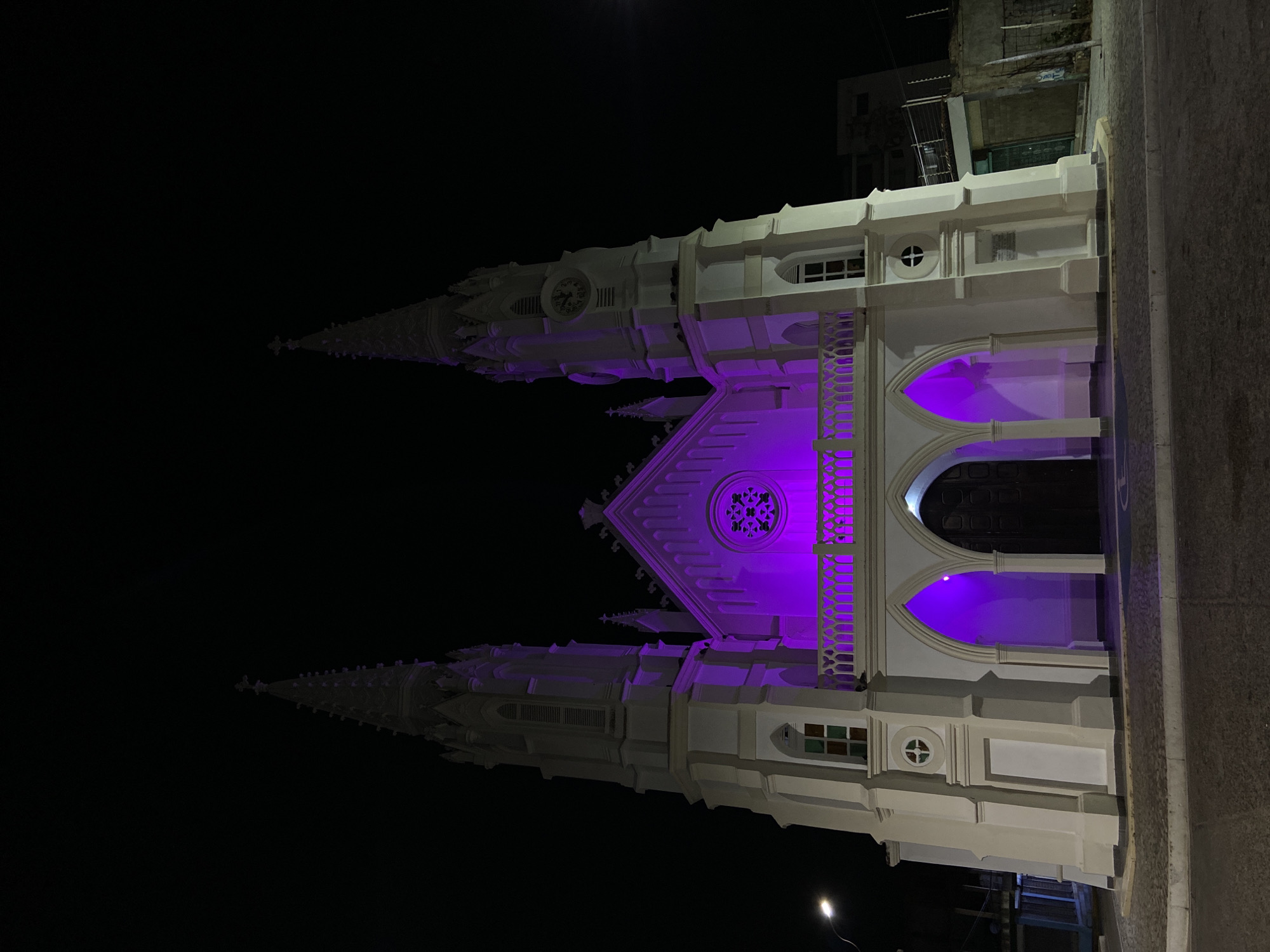 The image displays the exterior of the Iglesia San Juan Evangelista in Juan Griego, Venezuela, at night. The prominent Neo-Gothic style church features a white facade, dominated by two tall spires topped with crosses. Clock faces are visible on the upper sections of the spires. The central portion of the church, including a large rose window, an ornate balustraded balcony, and the interior of the three arched main entrances, is illuminated with purple light. The lower and outer sections of the structure are lit with white light. Dark wooden double doors are visible within the central archway. The church stands on a paved area with a cobblestone pattern in the foreground, adjacent to an asphalt road. To the right, less illuminated ancillary structures with fencing are partially visible. No individuals are present in the scene.