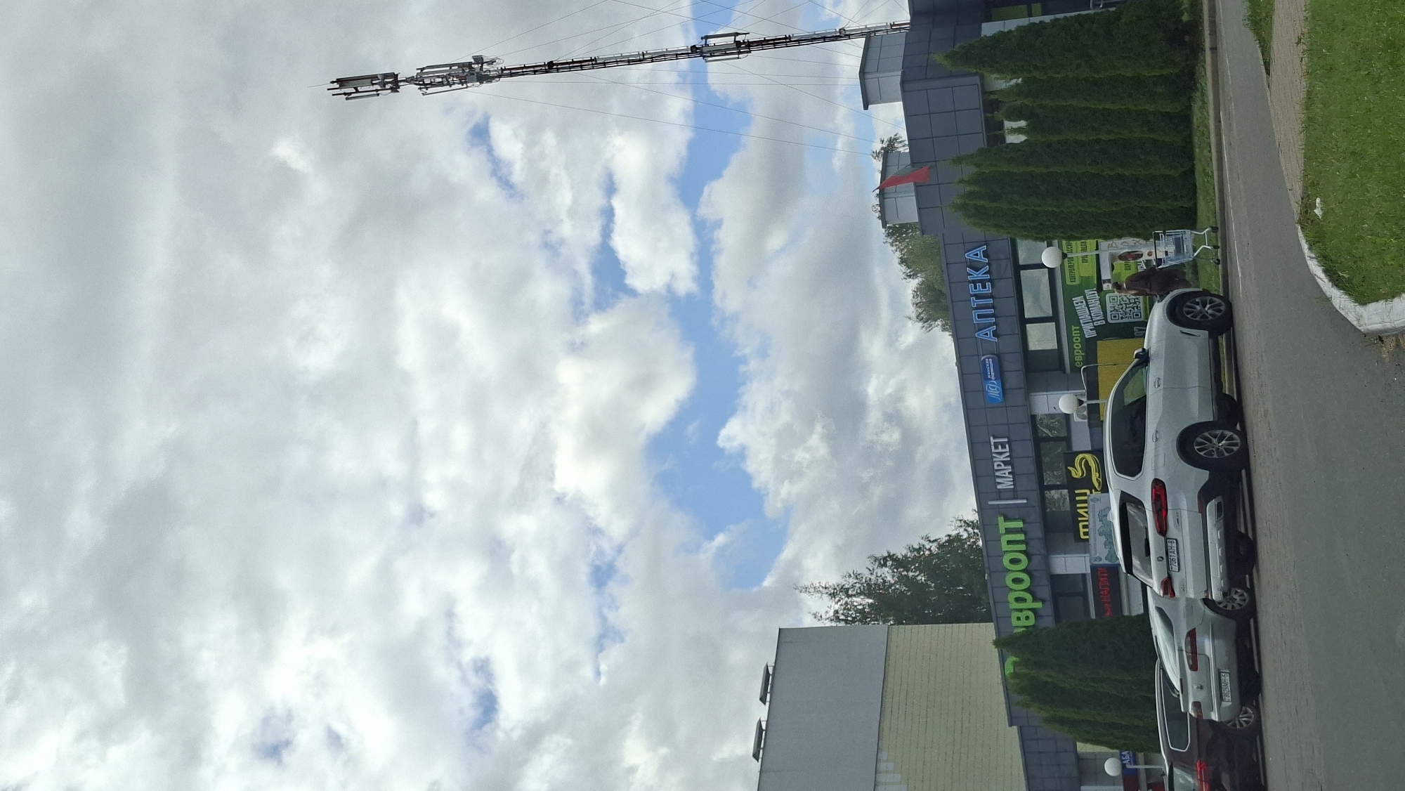 The image shows a street scene in Myadzyel, Belarus. A white BMW SUV is parked in the foreground on the right side of the image. Behind the SUV, there is a row of tall green trees. Beyond the trees is a building with a sign that reads "ЕВРООПТ МАРКЕТ" and "АПТЕКА" in Cyrillic. A person is walking to the right, pushing a shopping cart. In the background, a cell tower is visible against a cloudy sky. There are other cars parked in front of the building.