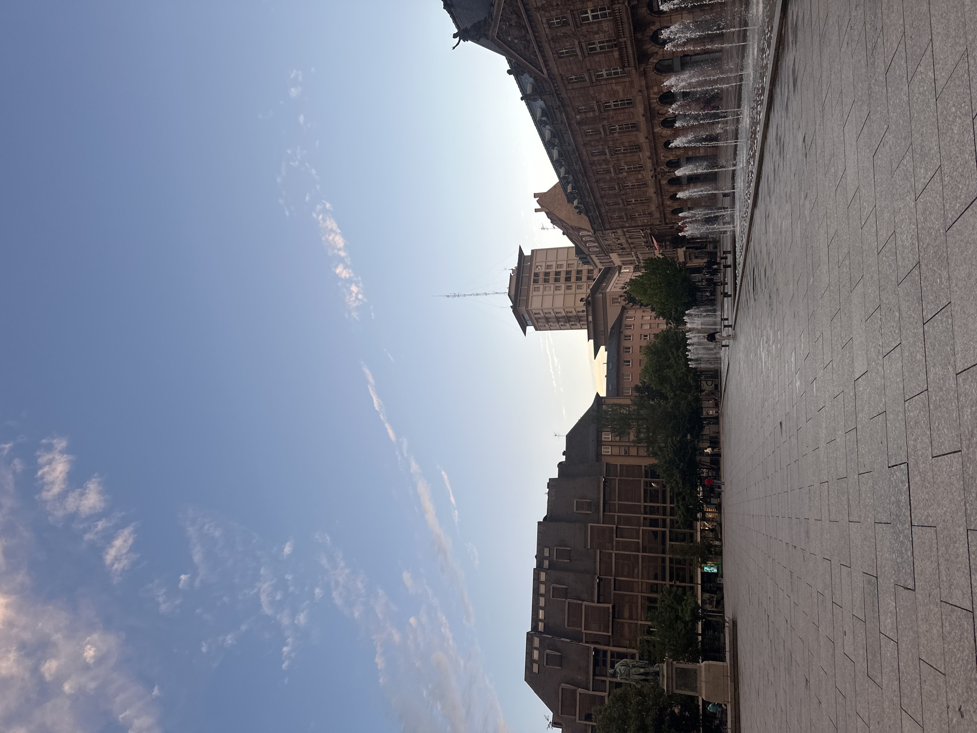 This image depicts Place Kléber, a central square in Strasbourg, France. The view showcases a wide, paved plaza with buildings on either side. A fountain runs along the edge of one building, creating a curtain of water. A modern-style building with a grid-like façade is visible on the other side. A tall building with an antenna stands in the background. The sky is clear blue with wisps of clouds. No people are clearly visible in the image.