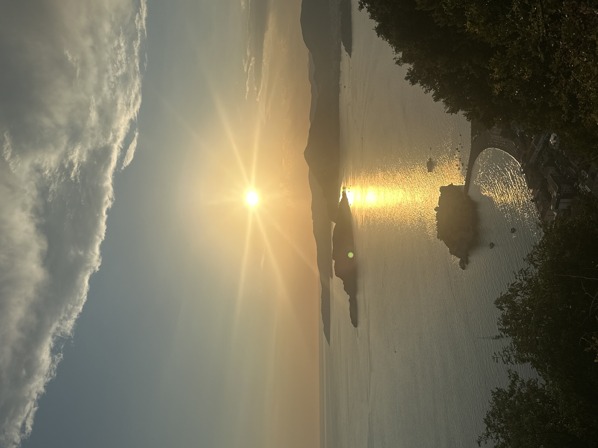 An aerial view captures the golden hour sunset over the Adriatic Sea, with Sveti Stefan, Montenegro, illuminated in the foreground. The sun, a bright, starburst-like orb, dominates the upper center of the frame, casting a shimmering golden reflection across the calm water. Jagged, silhouetted islands and peninsulas dot the horizon, their forms softened by the atmospheric haze.
In the mid-ground, the iconic islet of Sveti Stefan is clearly visible, connected to the mainland by a narrow causeway. The red-tiled roofs of the luxury resort are discernible, nestled amongst the island's architecture. A few boats are scattered in the water around the islet and further out at sea.
The foreground is partially obscured by the dense, dark green foliage of trees, suggesting the viewpoint is from a hillside overlooking the scene. The sky is a gradient of soft blues and yellows, with wisps of clouds catching the last rays of sunlight. The overall impression is one of serene beauty and tranquility, bathed in the warm glow of the setting sun. There are no discernible people in the image.
