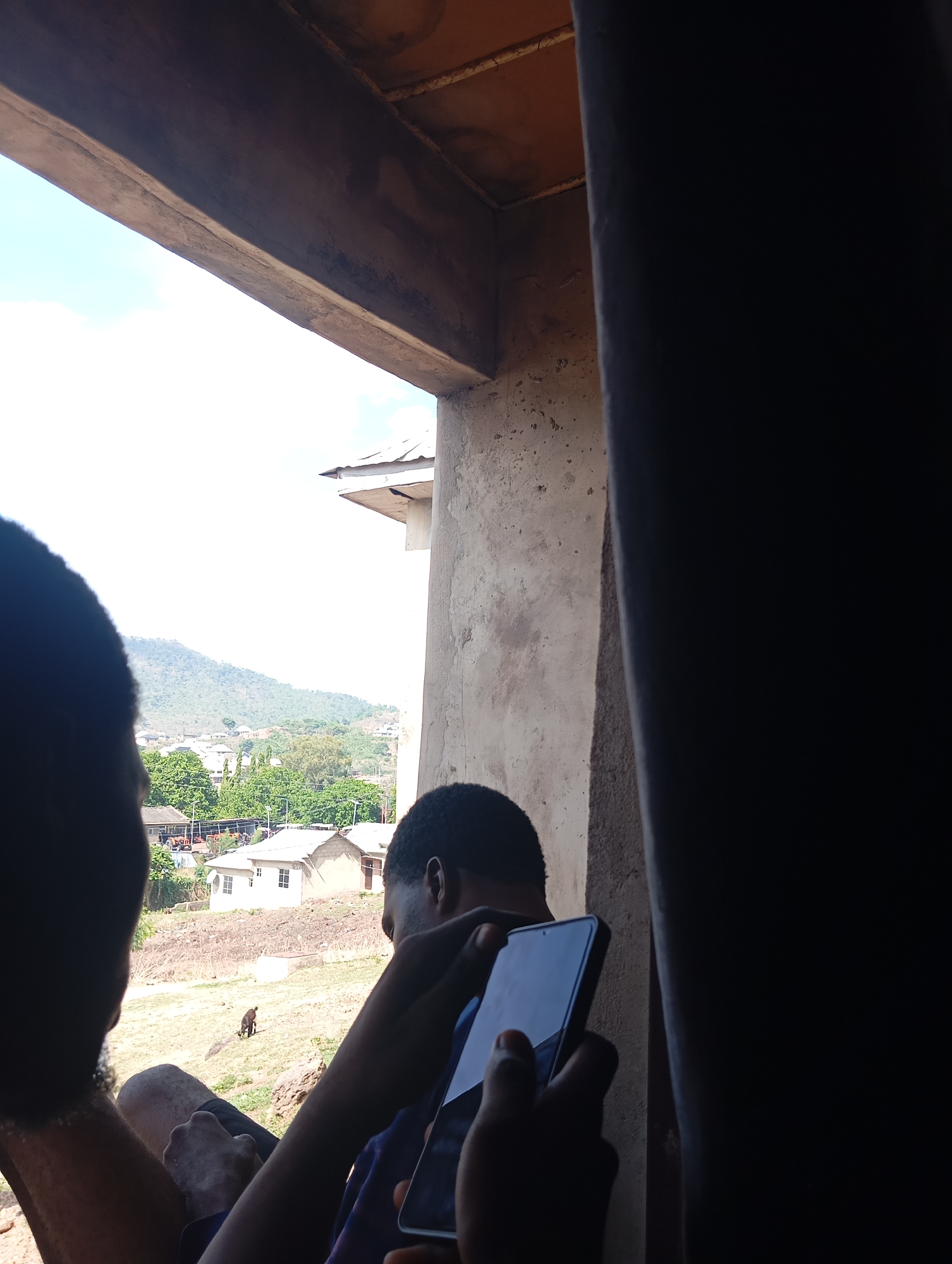 From an interior perspective, two people are viewing an outdoor scene through an opening. One is holding a black smartphone. In the background, houses are visible in the city of Lokoja, Nigeria. A hillside covered in trees is visible. A small animal is visible near the houses. The opening is surrounded by concrete structures.