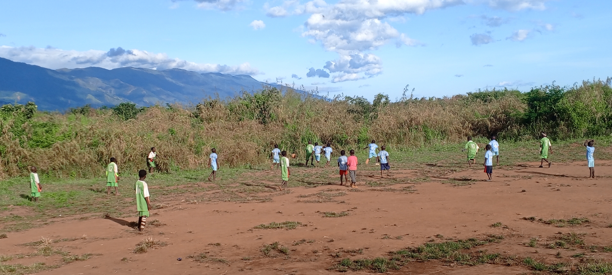 The image captures an outdoor scene in Ngalula, Democratic Republic of the Congo, depicting approximately twenty children, primarily boys, engaged in what appears to be a game of soccer. The setting is a large, open, reddish-brown dirt field with scattered patches of green grass in the foreground. In the midground, a dense band of tall, dry grass and green bushes forms a natural backdrop.
In the background, majestic, layered mountains rise under a bright blue sky dotted with scattered white clouds. The mountains display patches of green vegetation. The bright daylight, evidenced by relatively short shadows, suggests it is late morning or early afternoon, under clear and sunny weather conditions.
The children are actively spread across the field, running, standing, and moving as if in play. They are mostly clad in simple sports attire, forming at least two teams. One team wears light green jerseys with white trim and green shorts; visible numbers on their backs include '12', '10', and '9'. Another team wears light blue jerseys and blue shorts, with numbers like '9' and '10' visible on their backs. One child is also seen in a red shirt and blue shorts. Most children appear to be barefoot. A small, light-colored object, possibly a makeshift ball, is visible near some of the children. The scene evokes youthful energy and community engagement amidst a natural, rural landscape.