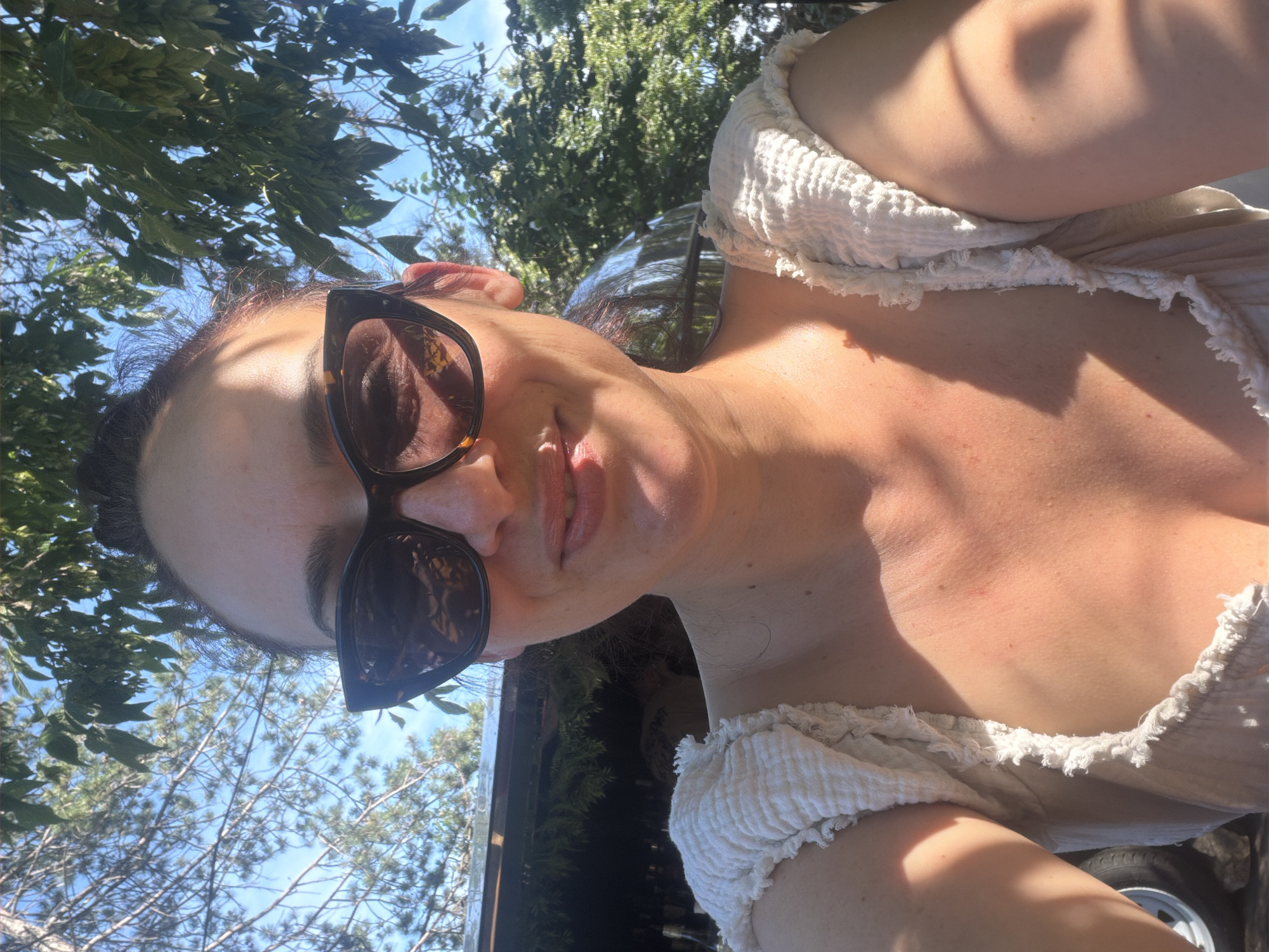 A woman is taking a selfie outdoors. She is wearing a white, ruffled top and large sunglasses with a tortoiseshell pattern. Her hair is pulled back. Sunlight is visible on her face and chest. Behind her, trees and a blue sky are visible. A portion of a vehicle and some bottles are also visible in the background. The location is not identifiable as Sozopol, Bulgaria, based on the provided image.