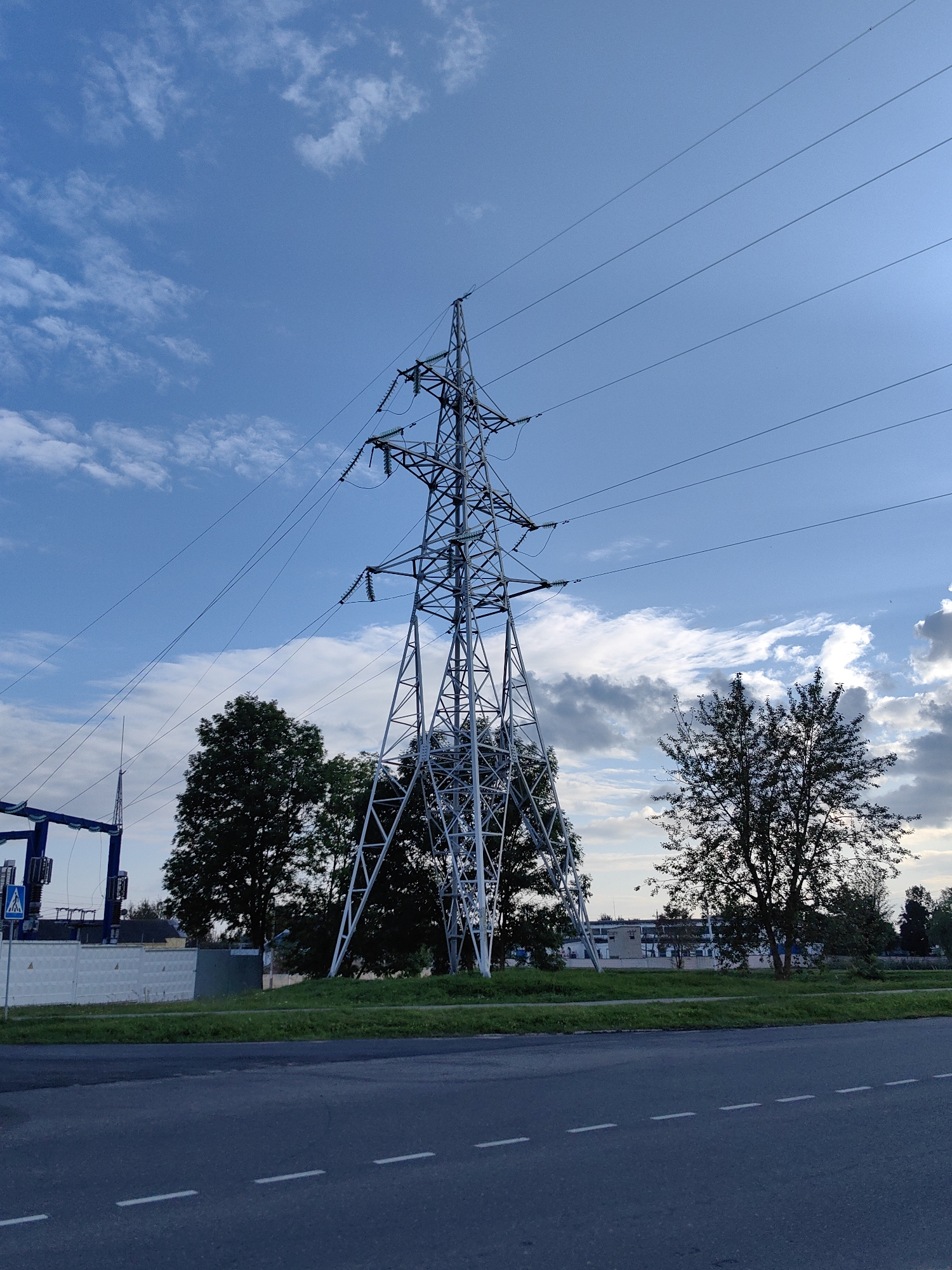 In Myadzyel, Belarus, a large electricity pylon stands prominently against a partially cloudy blue sky. The pylon, constructed of metal lattice, supports multiple power lines that stretch across the frame. In the foreground, a dark asphalt road with white dashed markings is visible. Behind the pylon, lush green grass lines the ground, separating it from a fence and a few trees. To the left, a section of a blue industrial structure with some equipment is partially visible, along with a pedestrian crossing sign. In the background, buildings and more trees can be seen under the expansive sky.