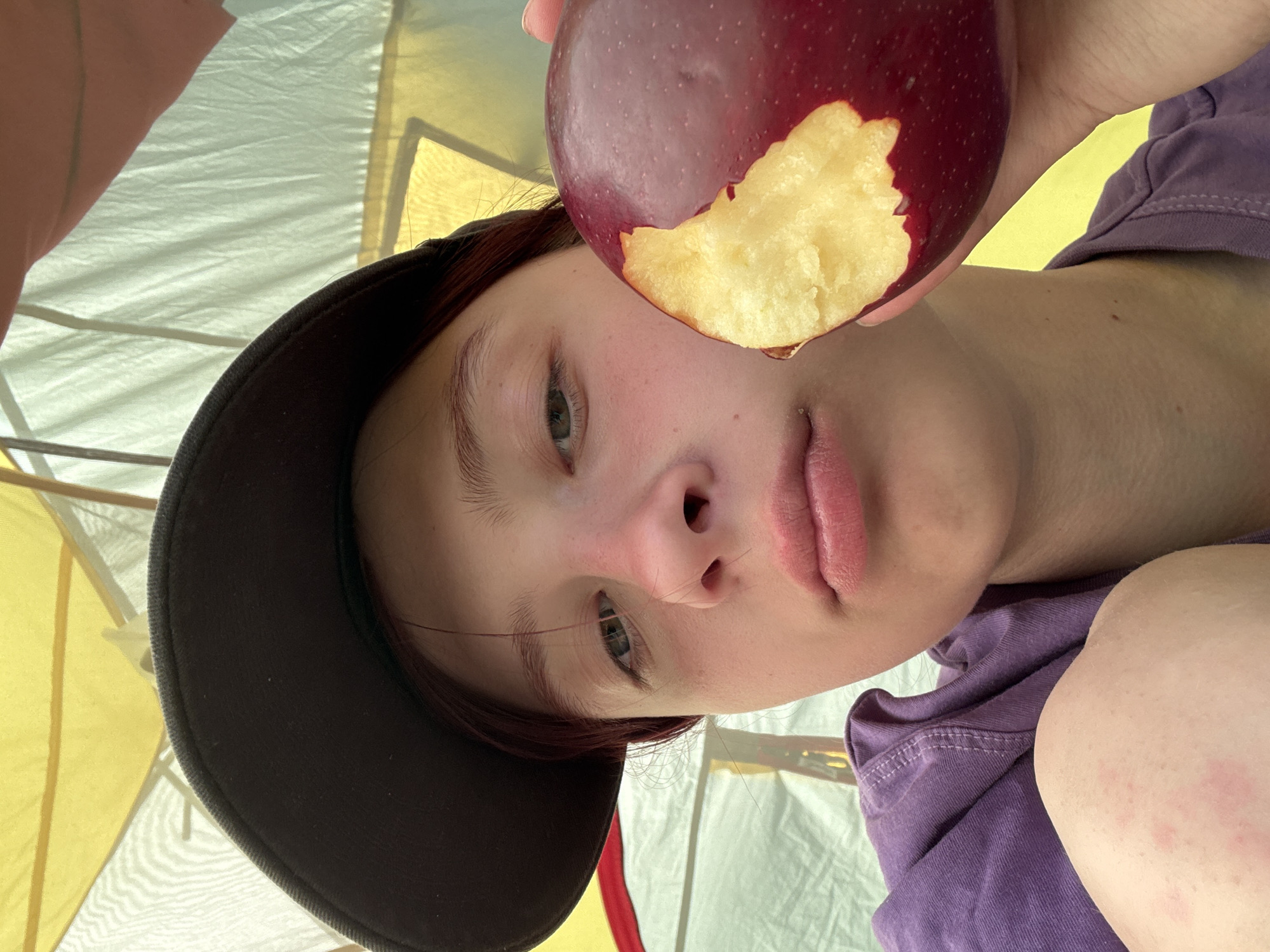 A person with fair skin, wearing a dark hat and a purple shirt, holds a partially eaten red apple in front of their face. The background is a tent-like structure with yellow and white panels. The person appears to be indoors. There is no information to indicate that the image was taken in Khvalynsk, Russia.