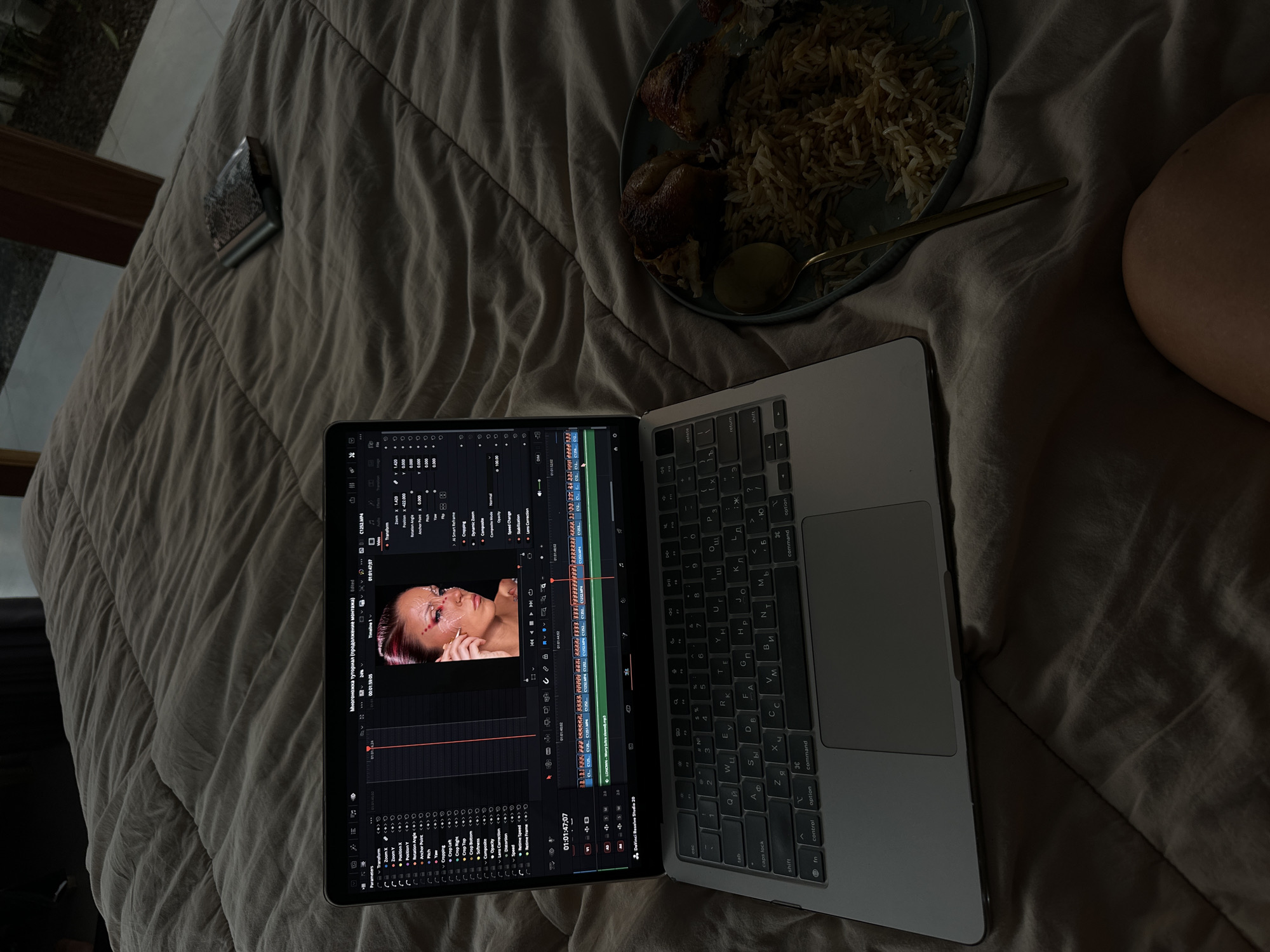 The image captures a scene on a bed in what might be a living or hotel space in North Kuta, Indonesia. A laptop sits open, displaying video editing software showing footage of a woman with pink hair and elaborate makeup. A plate of rice and dark meat is on the bed near the laptop. A small metallic object is on the bed to the left of the laptop. A person's leg is visible on the bed to the right of the laptop and food.