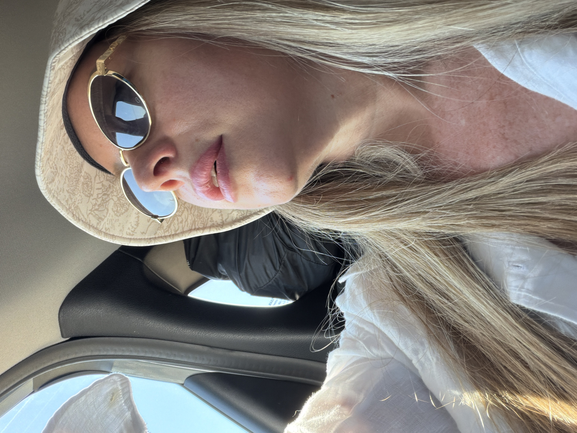 A medium shot captures a woman wearing a patterned bucket hat and gold-rimmed oval sunglasses from a low angle. She is looking upwards and to the right. Her lips are slightly parted, revealing her teeth. She has long, blonde hair and is wearing a white collared shirt. Sunlight illuminates her face and hair, creating a warm glow. The background is slightly blurred, showing the interior of a car. A black headrest is visible behind her, and the blue sky can be seen through the car window. The scene is likely within the city of Άγιοι Απόστολοι, Greece, given the context provided.
