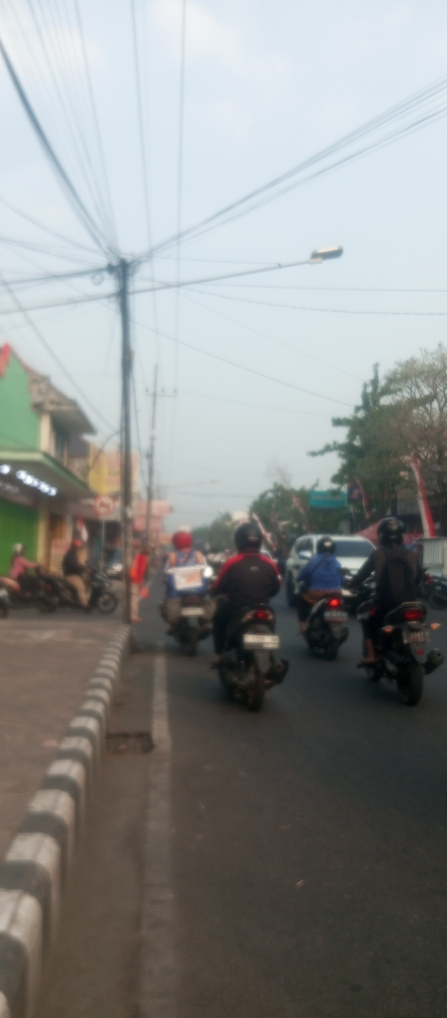 The image captures a street scene in Kediri City, Indonesia, likely on a hazy day given the muted colors and the overexposed sky. The foreground shows a paved road with a raised concrete curb on the left, marked with alternating black and white stripes. A significant portion of the left side of the image is dominated by a dense network of overhead electrical wires, with a utility pole visible.
On the road, several motorcycles are in motion. In the foreground on the right, a person wearing a black jacket with red accents is riding a black motorcycle with a license plate that appears to read "C 8928 BH". To the left of this motorcycle, another person in a blue jacket is riding a motorcycle. Further ahead and to the left, a person in a red helmet and a backpack is on a motorcycle, followed by another motorcycle with a rider in a red helmet. On the far left, a few motorcycles are parked along the side of the road, with a person in pink attire on one of them, near a green building with a storefront.
The background features more buildings, some with red accents, and lush green trees. There are also some red and white flags or banners visible on poles. A white car is partially visible on the road. The overall atmosphere is that of a typical urban street with moderate traffic, characterized by the prevalence of motorcycles.