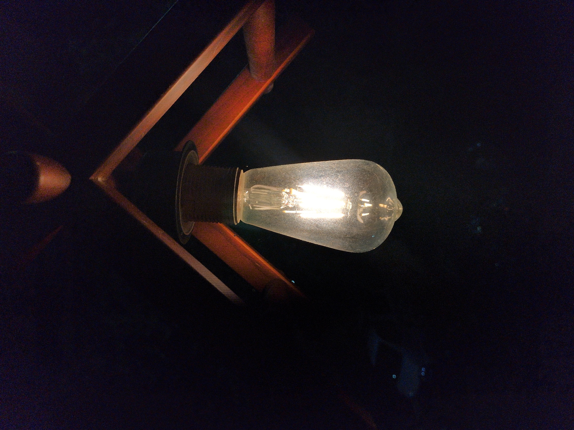 A close-up, low-light shot shows an illuminated Edison bulb hanging from a dark fixture. The bulb, with its visible filament, is the central subject. Behind the bulb, the dark background is partially obscured by angled, reddish-brown wooden beams, part of the fixture's construction. The overall ambiance is dark and moody, with some faint light reflections visible in the lower left quadrant, possibly from distant objects or light sources in Bhagsu, India.