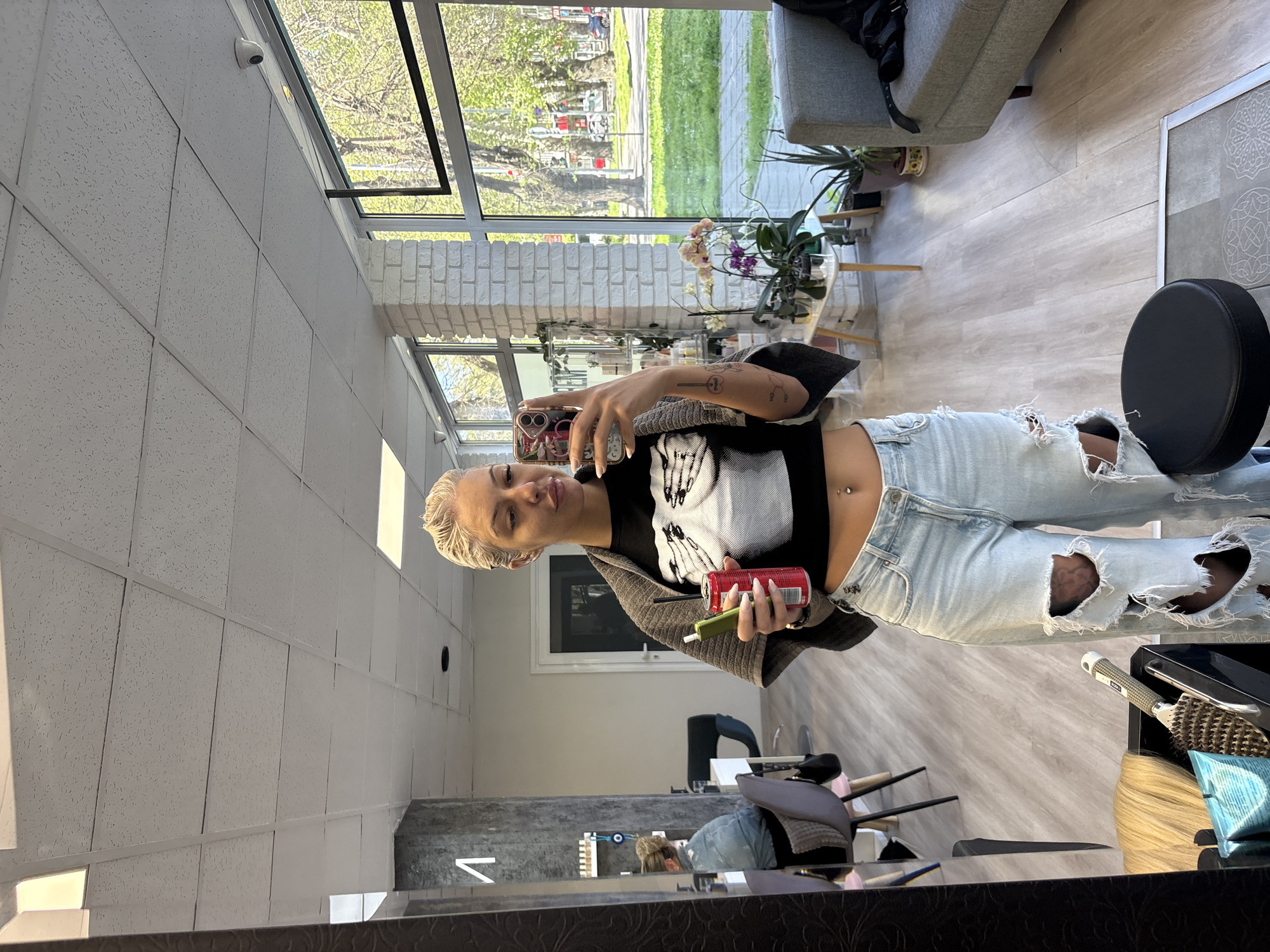 The image, taken in Plovdiv, Bulgaria, captures a mirror selfie of a young woman in what appears to be a modern hair salon or beauty studio. The image is oriented vertically (portrait mode), but the woman is depicted standing upright, looking into the mirror with her phone.
The primary subject is a young woman with short, platinum blonde hair. She has a nose ring and a navel piercing, and visible tattoos on her left arm. She is dressed in a black cropped t-shirt featuring a white, distressed graphic design, a grey open cardigan, and light-wash ripped jeans with large tears. In her left hand, she holds a red soda can (identifiable as Coca-Cola), and in her right, a green vape pen. Her expression is neutral as she looks into the mirrored phone. In the lower part of the mirror's reflection, another person, possibly a salon employee, is visible looking downwards.
The setting is an indoor commercial space, likely a salon, characterized by bright natural light. The room features light wood-look flooring and white walls, with a white drop-tile ceiling. Large windows line one wall, offering a view of a sunny street with green trees and grass, confirming it is daytime. A section of white exposed brick adds a decorative element. Furnishings include a grey sofa or armchair with a black bag resting on it, and a small glass table adorned with potted orchid plants and other greenery. To the right, a black round cushion or stool is visible, along with a counter displaying salon tools like brushes and what appears to be blonde hair extensions in a box.
The activity depicted is the woman taking a mirror selfie, suggesting she is relaxing or waiting in the salon. Notable details include the "Coca-Cola" text on the can and a 'V' shape visible in the mirror's reflection on a wall, potentially part of a logo or sign. The overall ambiance is bright and contemporary.