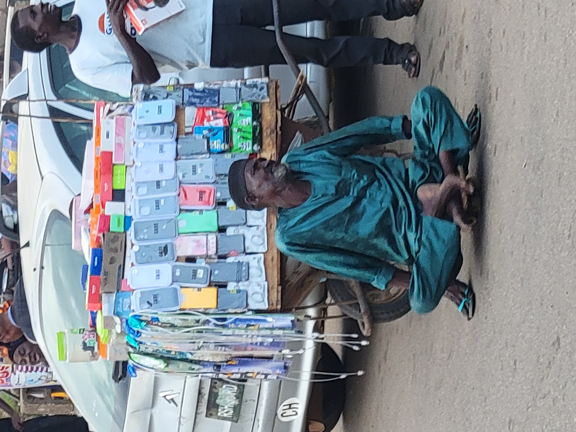 In Barakin Lerawi, Nigeria, a man in a teal traditional outfit sits cross-legged on the asphalt ground next to a cart laden with phone cases and accessories. The cart is positioned near the back of a silver car. The man, with a weathered face and grey beard, is looking to his right. A second man, wearing a white t-shirt with a red and orange logo and dark trousers, stands to the right of the cart, holding an orange box. The background shows a blurred street scene with other people and a portion of a white car. The ground is a rough, grey asphalt surface. The overall scene depicts a street vendor setup.