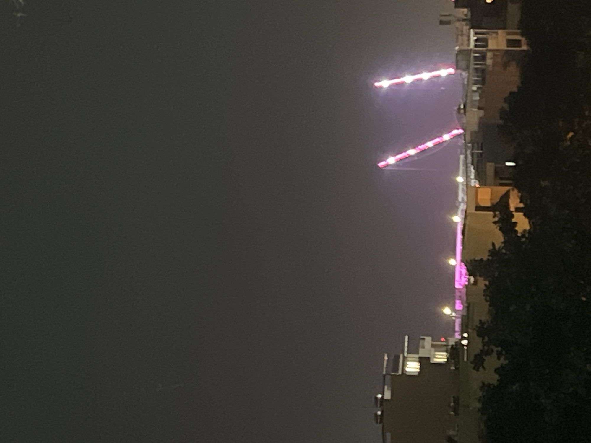 The image depicts a nighttime city scene in Đà Nẵng, Vietnam, dominated by a dark, hazy sky and the silhouettes of buildings and trees. In the mid-ground, two large construction cranes, illuminated with pink and white lights, extend upwards. Their forms suggest they are actively involved in a construction project. Below and to the left of the cranes, several buildings are visible, some with illuminated windows, indicating occupancy. The foreground is obscured by dark foliage, likely trees, suggesting the vantage point is from a higher elevation or across a green space. The overall impression is one of urban development and activity occurring under the cover of night.