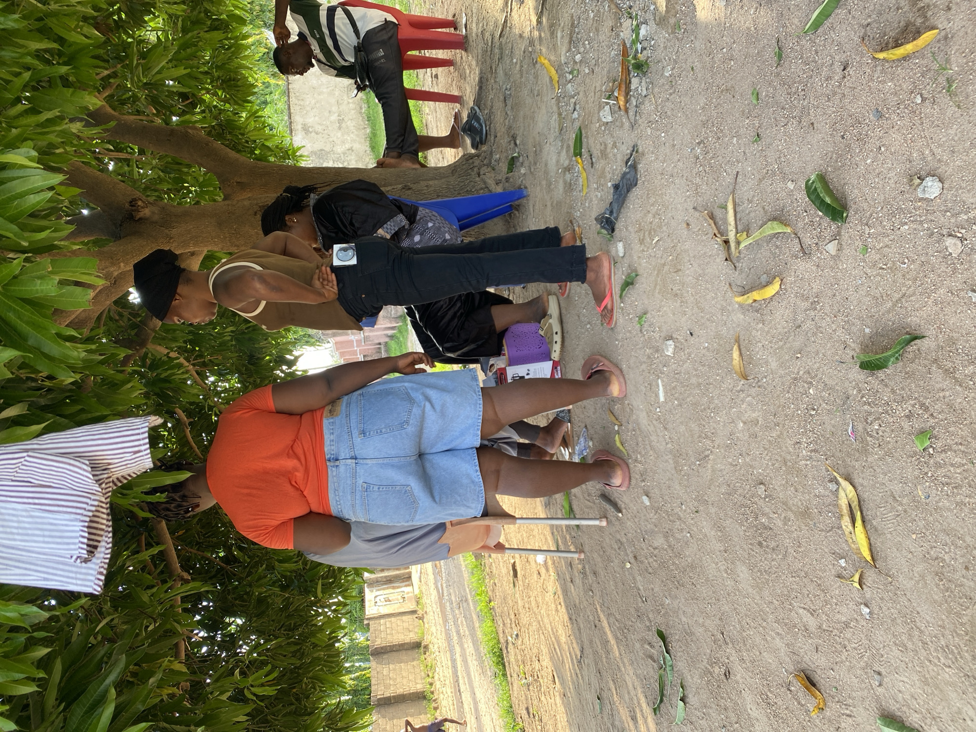 In Howayi, Nigeria, three women and one man are gathered under a tree. One woman, wearing an orange t-shirt and denim shorts, stands facing away from the camera. To her left, a second woman in a brown tank top and black pants is also facing away. A third woman, also in black, is partially visible behind them. The man, wearing a striped shirt and black pants, sits on a red chair to the right of the frame, looking at his phone. Fallen leaves are scattered on the sandy ground around them, and a piece of striped fabric hangs from a branch above. The environment appears to be an outdoor area, possibly a yard or public space, with a brick wall visible in the background.