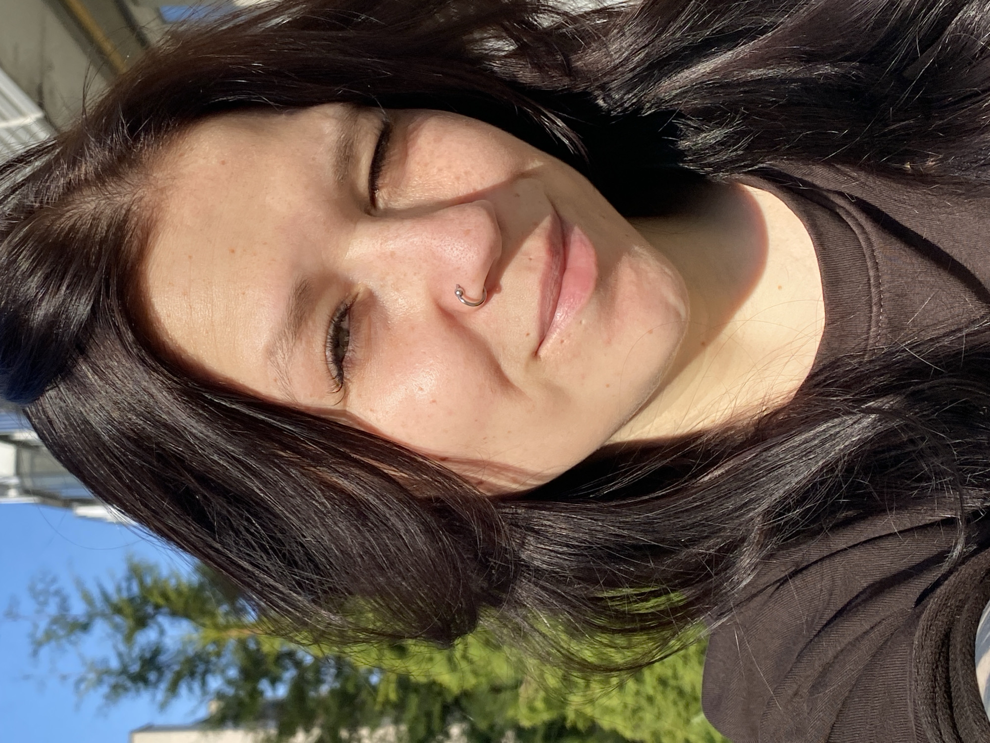The image is a close-up selfie of a woman with dark hair, freckles, and a nose ring. Her eyes are closed, and she has a slight smile. She's wearing a dark-colored shirt. The background shows a blue sky, green trees, and part of a building. There's no definitive information in the image to pinpoint the location as Chaikovsky, Russia. The location is unconfirmed based solely on the image.