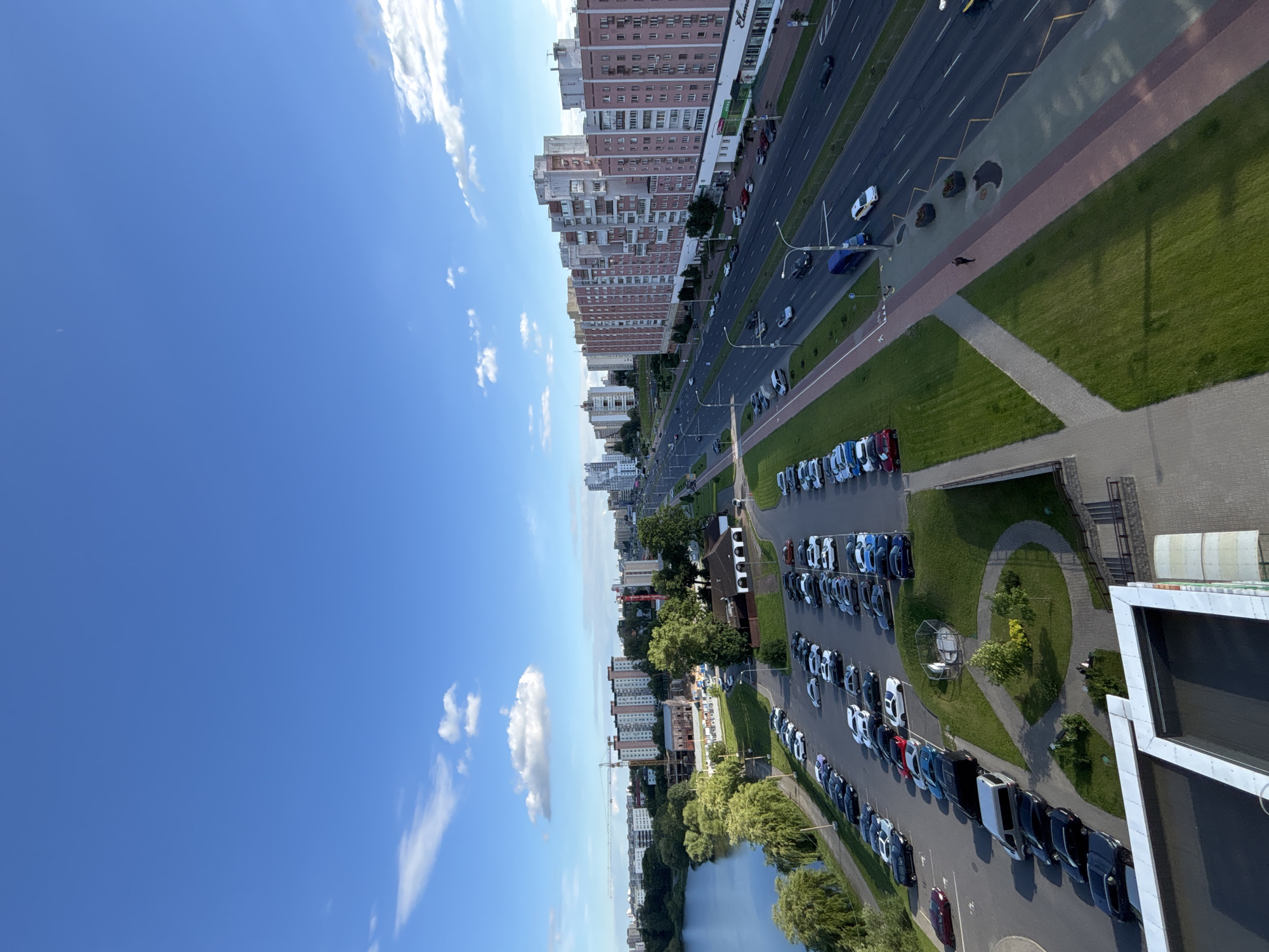 The image captures a high-angle view of a cityscape in Minsk, Belarus, dominated by a wide, multi-lane road and a body of water. The scene is set on a sunny day with scattered clouds, indicated by the bright blue sky and distinct white clouds.
On the left side of the frame, a calm body of water reflects the sky, bordered by lush green trees and vegetation. Beyond the water, residential buildings with varying architectural styles and heights are visible, some appearing to be under construction, as suggested by the presence of a crane.
The main focus of the right side of the image is a busy road with several lanes of traffic. Cars of various colors are driving and parked along the roadside. Adjacent to the road are green grassy areas and pedestrian walkways. A significant portion of the image is dedicated to a large parking lot filled with many cars, predominantly black, white, and blue.
The perspective suggests the photo was taken from a high vantage point, possibly a tall building, looking down and across the urban landscape. The time of day appears to be daytime, judging by the strong sunlight and clear visibility. There is no visible text in the image. The overall impression is one of a typical urban environment with a mix of residential, commercial, and recreational spaces.