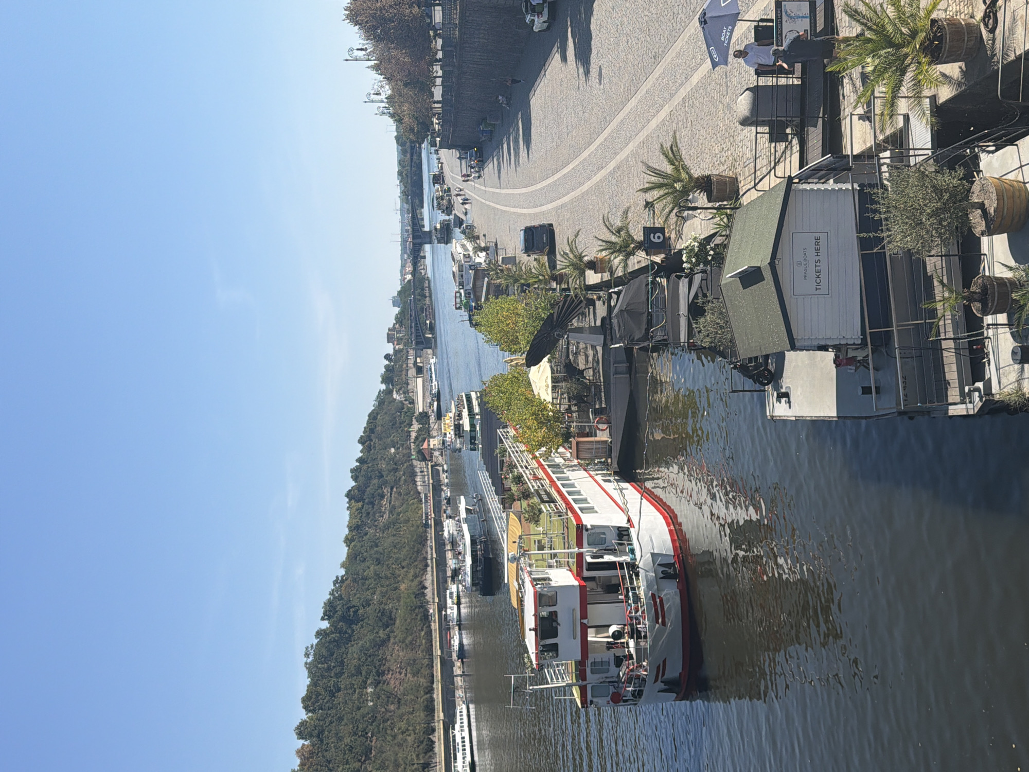 The image depicts a sunny day along the Vltava River in Prague, Czechia. In the foreground, a large, white and red tour boat is docked. The boat has multiple decks, some with greenery and seating. Adjacent to the boat is a floating ticket booth with the sign "PRAGUE BOATS TICKETS HERE." Further along the riverbank, a cobblestone promenade is visible, with several other boats docked and a row of lampposts. People are seen walking and standing on the promenade. In the background, bridges span the river, and a tree-covered hillside rises on the far bank. The sky is clear and blue.