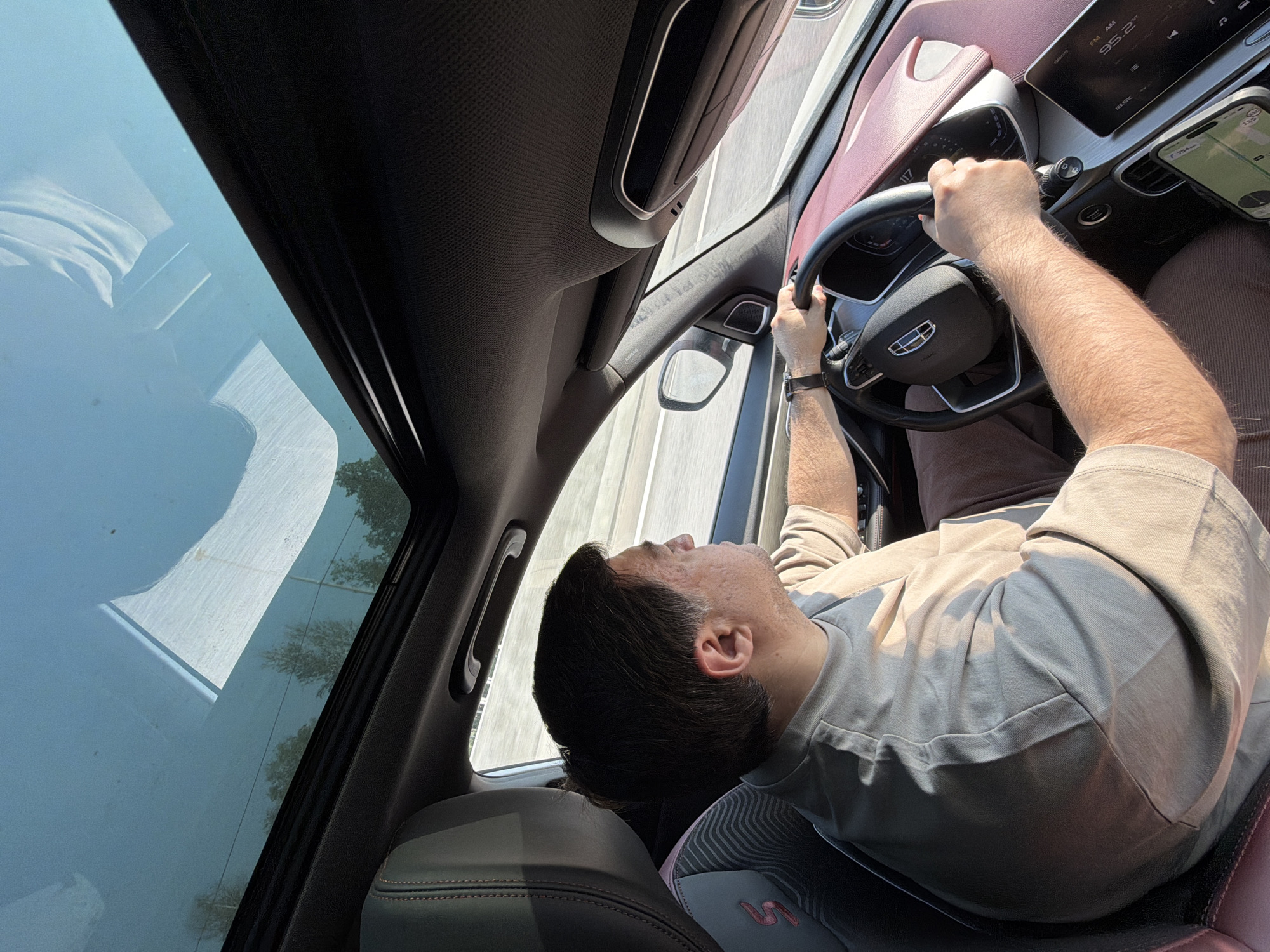 In the city of Fyodorovo, Russia, a man is driving a car with a panoramic sunroof open. He is wearing a light-colored t-shirt and brown pants. The interior of the car is visible, including the steering wheel, dashboard, and seats. The sunroof allows a view of the sky and trees.