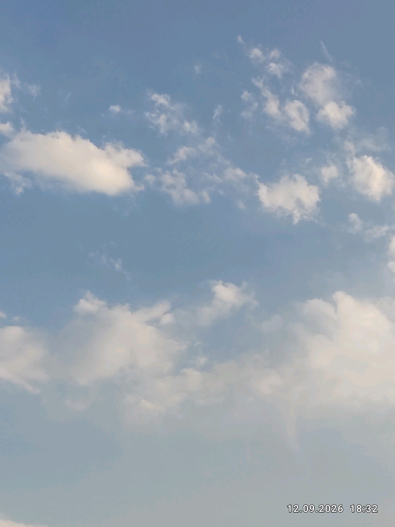 A clear, pale blue sky with scattered white cumulus clouds is depicted. The clouds vary in density and shape, with some appearing thin and wispy while others are more substantial and rounded. In the bottom right corner of the image, a timestamp reads "12.09.2026 18:32." No people, specific structures, or identifiable landmarks within Bishkek City, Kyrgyzstan, are visible in this sky-focused image. The environment is exclusively the sky and clouds.