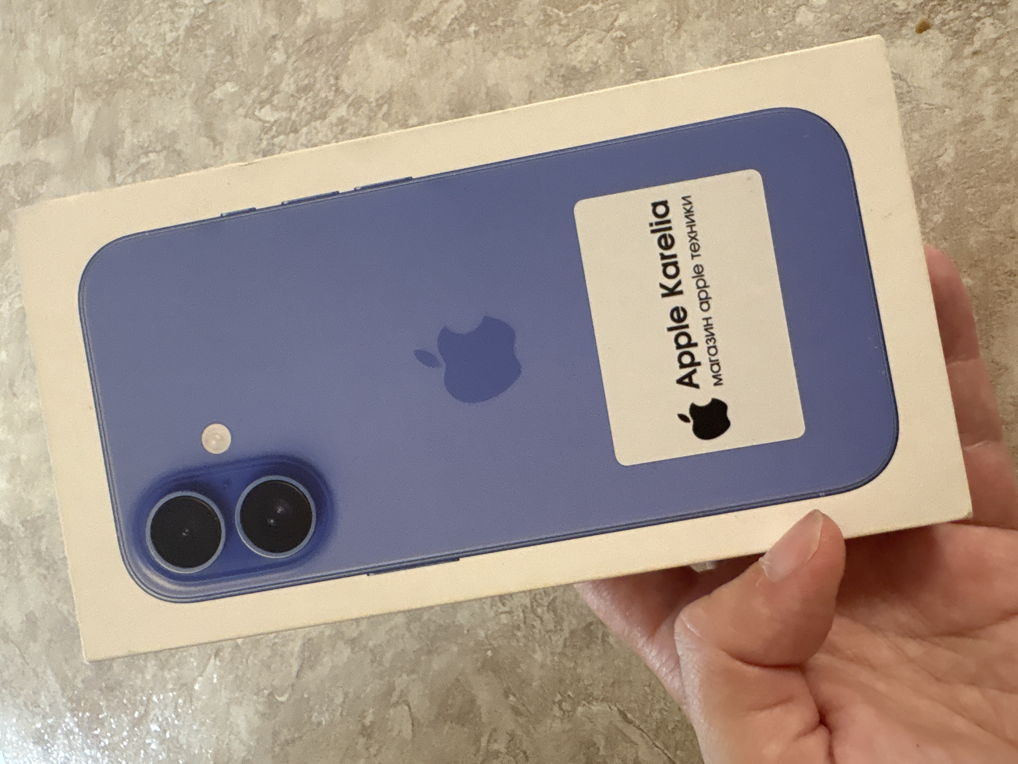 A person's hand is holding the box of an iPhone 12 in the color purple. The box is light beige with the Apple logo and text that says "Apple Karelia" and "магазин apple техники." The background is a speckled, light-brown surface. The image was taken on July 26, 2023, in Vetluzhsky, Russia.