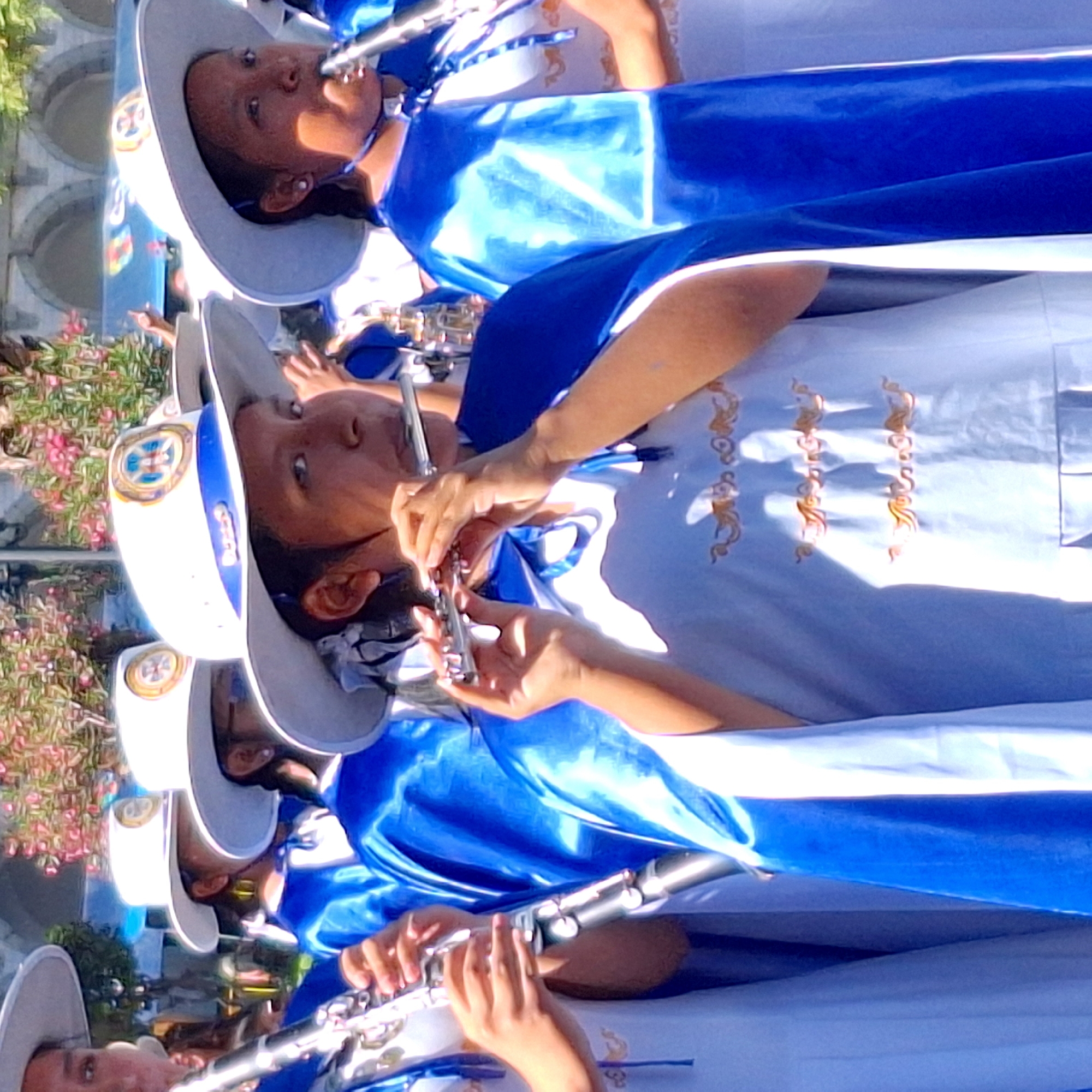 A vertical, medium-shot shows a group of people dressed in white and blue uniforms playing musical instruments. The location appears to be a city, though the specific details of the location remain unknown. The people in the group are all wearing white hats with blue trim and a circular emblem on the front. They are also wearing white shirts with gold embellishments, and blue capes with white trim. The people in the group are playing instruments, including flutes and clarinets. The instruments are silver in color, and the people are holding them with both hands. The image is well-lit and in focus, and the colors are vibrant.