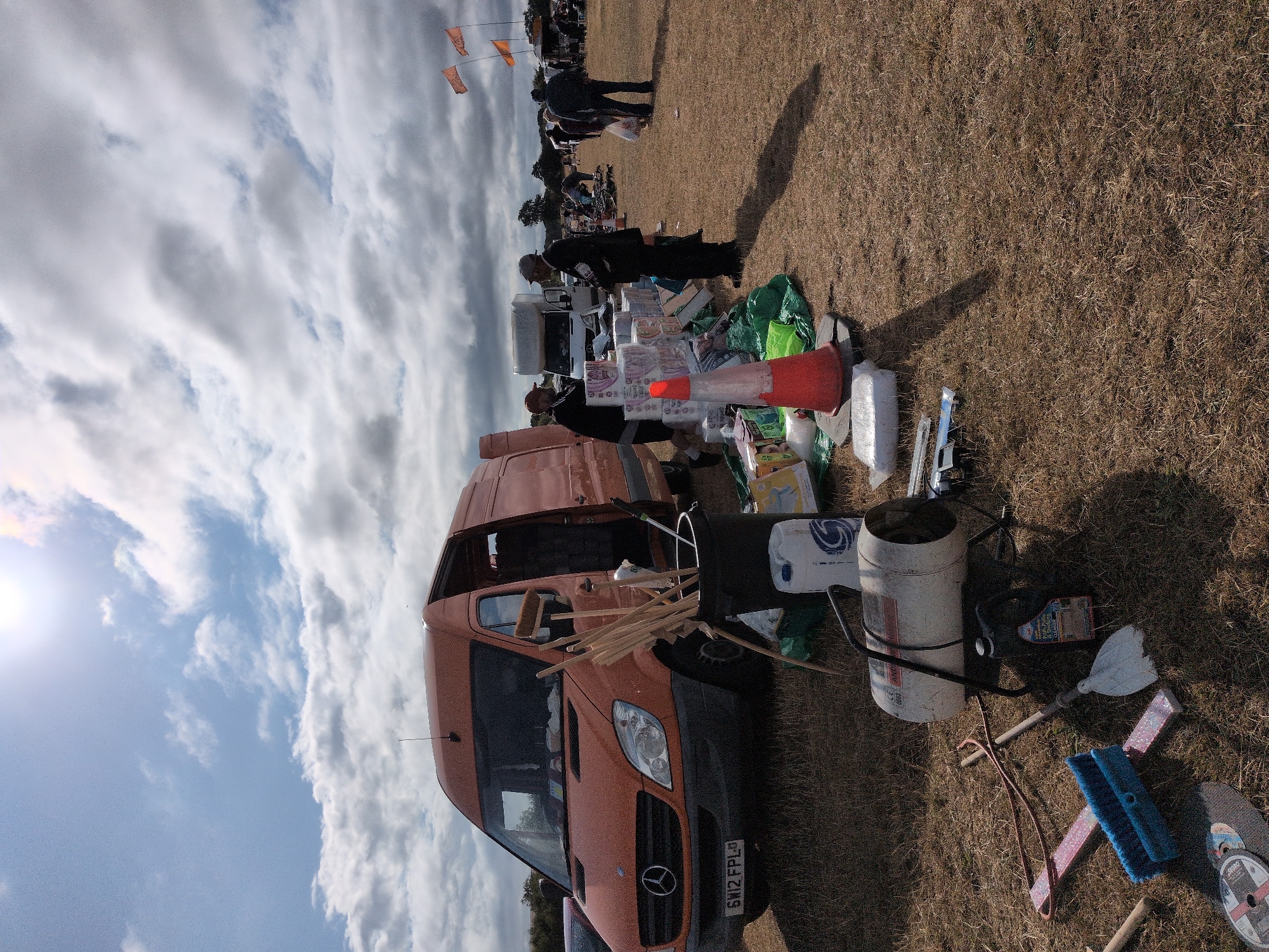 The image depicts an outdoor market scene, likely a car boot sale, on a sunny day with a partly cloudy sky. The foreground features various items laid out on the dry, grassy ground. These include a blue broom, a white dustpan, a metal object with a red hose attached, a circular metal saw blade, and a white cylindrical object with black handles and markings, possibly a heater or generator. A black oil container with a yellow label is also visible.
Dominating the mid-ground is an orange Mercedes-Benz Sprinter van with its driver's side door open. It has a license plate reading "SW12 FPL". Next to the van, a black bucket holds several wooden poles, suggesting some kind of construction or market stall setup. Piles of what appear to be toilet paper rolls are stacked to the left of the van. Two individuals are standing near these items; one is wearing a dark jacket and cap, and the other is wearing a dark jacket and a red cap.
Further into the background, another white van is parked. More market stalls and people are visible in the distance, with orange flags flying in the sky. The overall impression is a busy, casual market event taking place on a large, open field. The location is specified as South Staffordshire, United Kingdom.