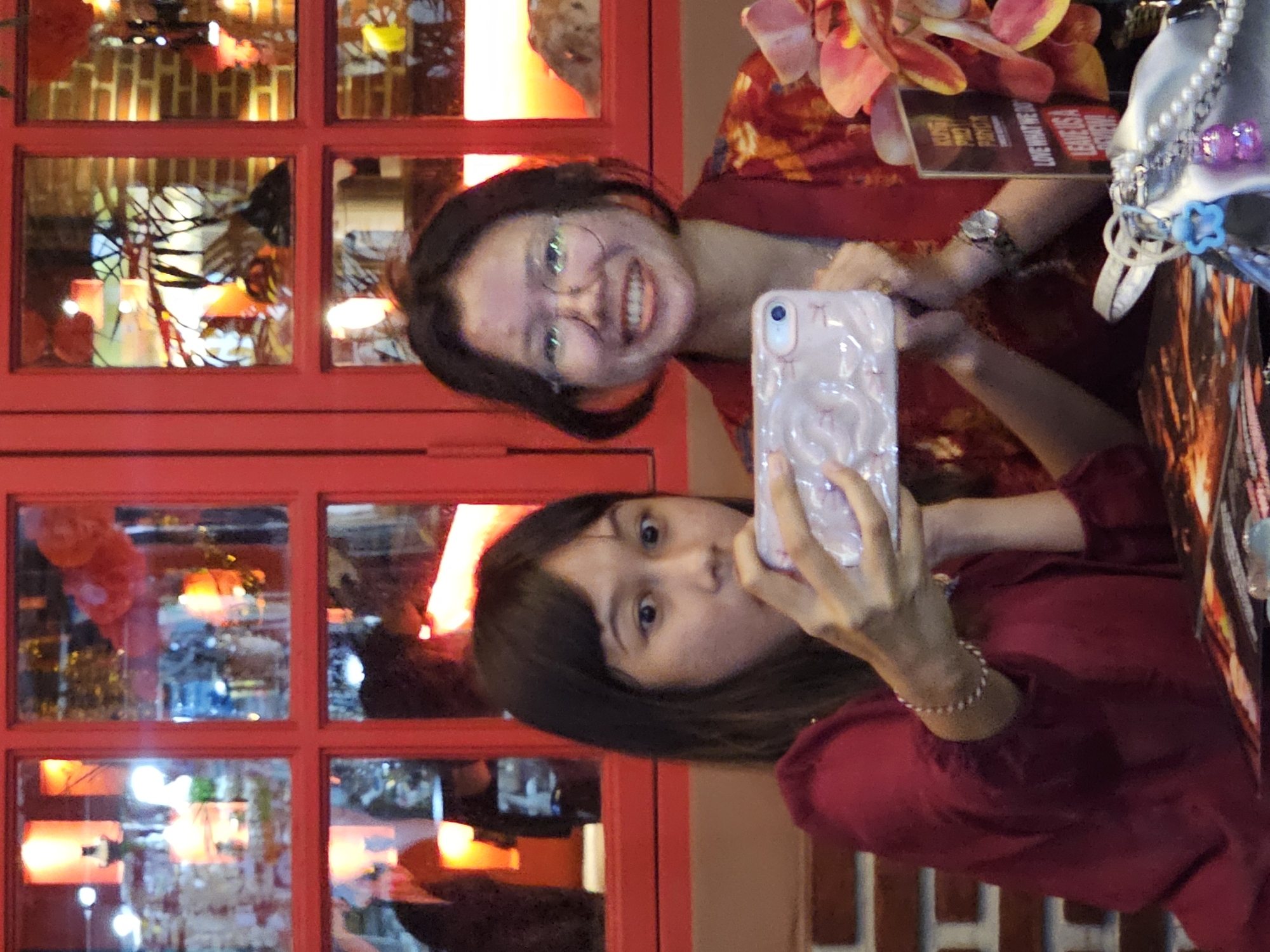 Two women are posing for a selfie in front of a red-framed window with multiple panes. The woman on the left, facing the camera, is holding a phone with a decorative case to take the picture. She has dark hair, bangs, and is wearing a red top. The woman on the right is smiling and also looking at the camera. She is wearing glasses and a red patterned top. In the background, reflections in the window panes suggest an interior setting with decorative lighting. Other objects visible include a purse with beads, flowers, and a menu. Based on the style of the red window frame and the overall ambiance, the location is likely a restaurant or establishment in the city of Kuta, Indonesia.