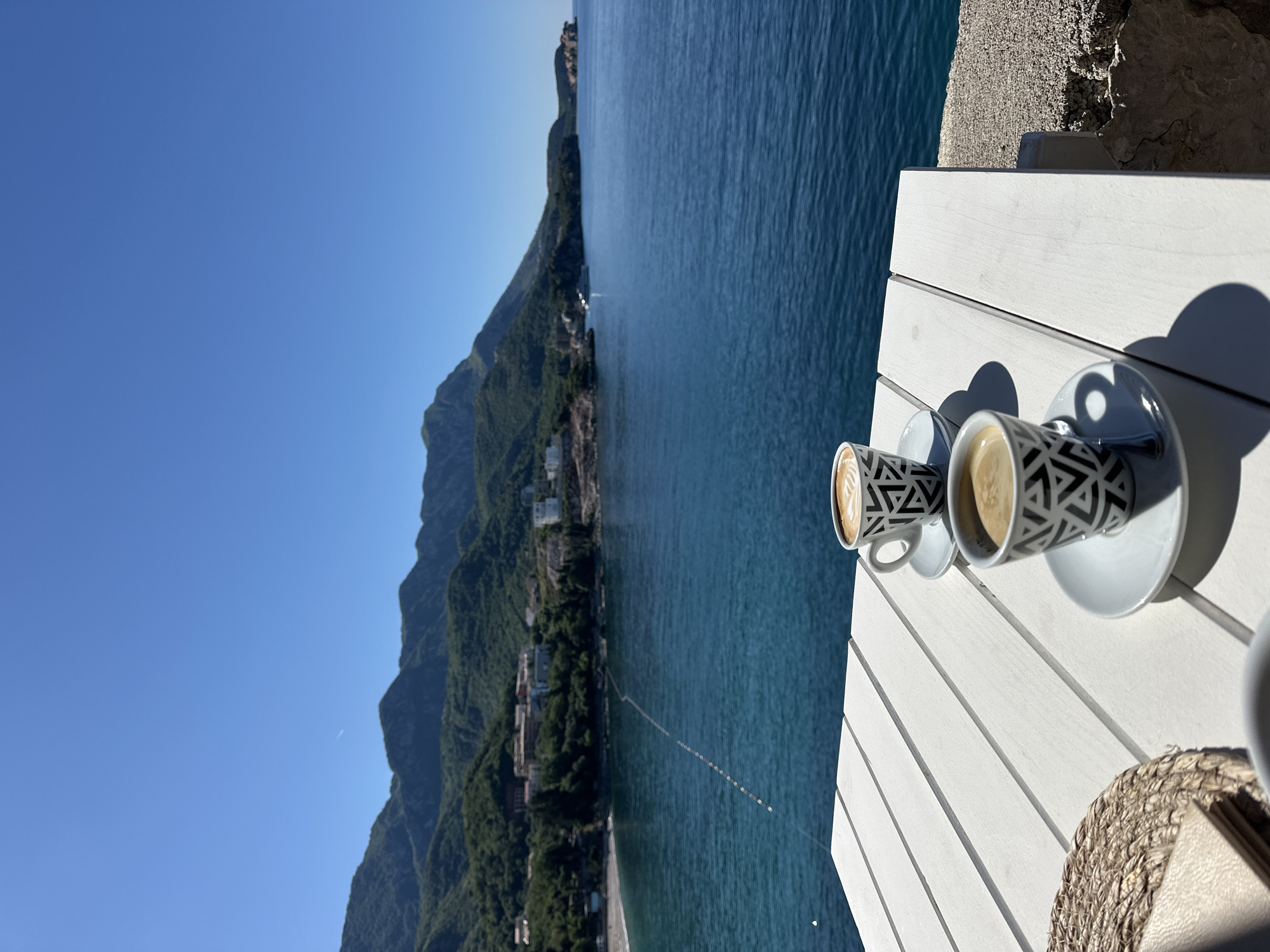 A high-angle, medium shot shows two cups of coffee on a white, slatted table on the edge of what appears to be a terrace. The cups are white with a black geometric pattern and are sitting on white saucers. The cups cast strong shadows. The backdrop reveals a scenic view of Boreti, Montenegro, featuring mountains covered in green vegetation, a clear blue sea, and a cloudless blue sky. A small beach with buildings is visible along the coastline. The scene suggests a peaceful and leisurely atmosphere.