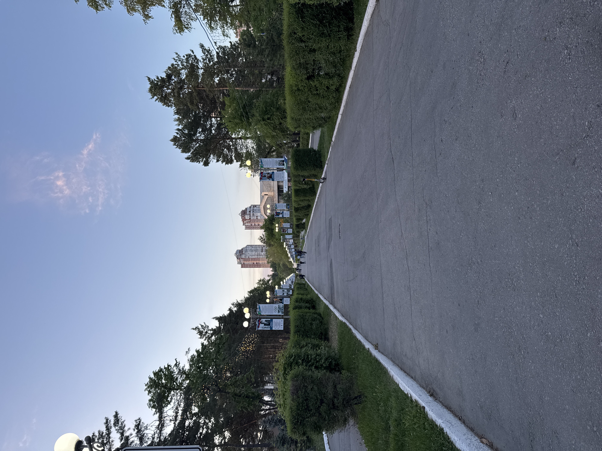 The image depicts a wide, asphalt pedestrian walkway or road in Khabarovsk, Russia. The walkway has a white painted curb along its right edge. Along the left side of the walkway, there is a row of trimmed bushes and trees, interspersed with streetlights that feature spherical lamps and what appear to be banners or signs. In the distance, several tall buildings can be seen. The sky is a clear blue with a few wispy clouds. The photo is taken at an angle, giving the impression that the walkway slopes upwards towards the buildings in the distance. The time of day appears to be late afternoon or early evening. There are distant figures walking on the walkway.