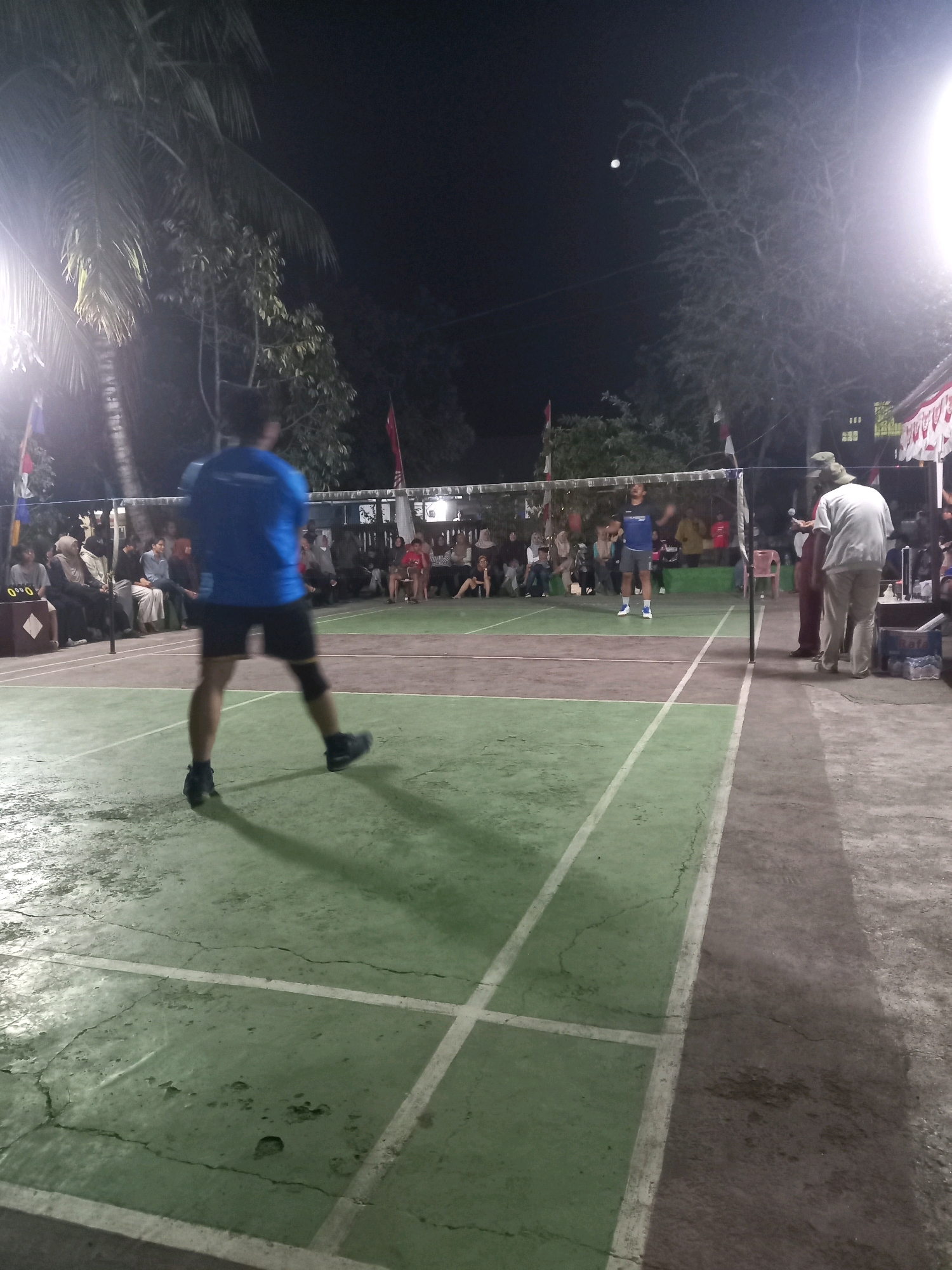 The image depicts an outdoor badminton court at night in Banyumulek, Indonesia, illuminated by artificial lights. In the foreground, a person wearing a blue t-shirt and black shorts is actively playing badminton, positioned in the center of the court, facing away from the camera. The court surface is green with white lines marking the boundaries. Spectators are seated on benches or chairs on the side of the court, mostly blurred due to the low light conditions. Trees, including a palm tree on the left, surround the court. A badminton net divides the court, and another player is visible on the opposite side. The background shows a dark sky with a bright object that could be the moon. The scene suggests a local sports event or casual game taking place after dark.