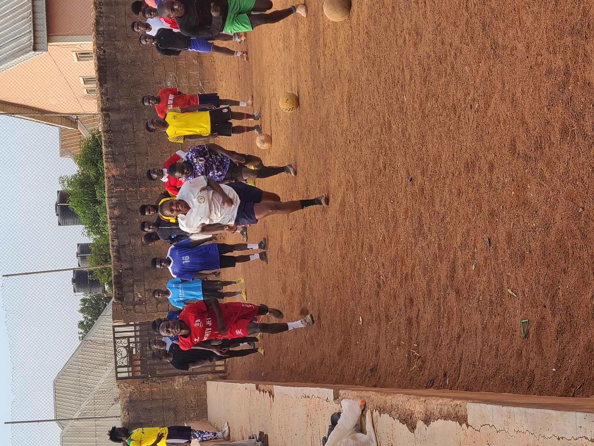 The image shows a group of approximately 15 young men, possibly soccer players, practicing on a dirt field in an urban or semi-urban area, location Unknown, Unknown. The men are wearing various sports jerseys and shorts. Three soccer balls are visible on the field. A brick wall runs along the side of the field, with buildings and water tanks visible in the background. A chain-link fence partially obscures the view on the left side of the image. The ground is a reddish-brown dirt surface. The sky is pale, indicating possible early morning or late afternoon light. Some of the men are in motion, seemingly kicking or interacting with the soccer balls.