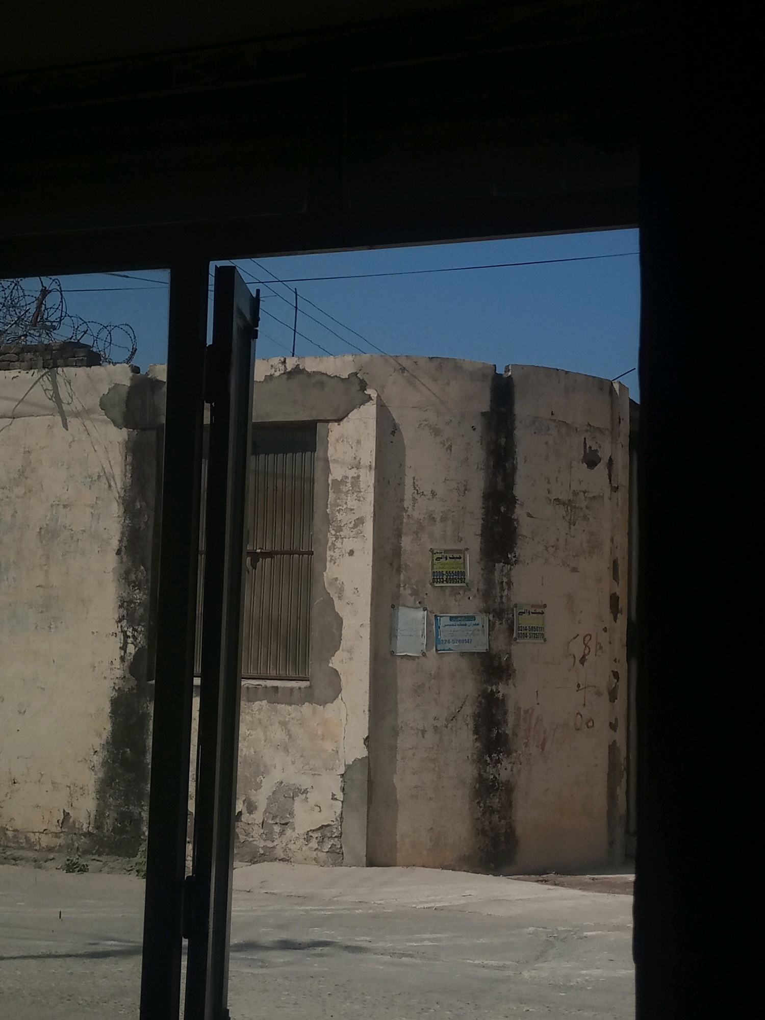 The image shows a view through a dark opening, possibly a doorway or window frame, into an outdoor area. In the background is a curved, weathered concrete structure, appearing to be a water tank or similar utility building. The wall of this structure is stained and peeling. On the wall, there are several posters or notices, some with Arabic script and phone numbers. To the left of the structure is a low concrete wall topped with coiled barbed wire. A metal-grated door is visible on the front of the concrete structure, partially obscured by vertical dark posts in the foreground, which are likely part of the opening through which the image is taken. The ground in front of the structure is paved with concrete, with some small weeds growing at the base of the wall. The sky above is a clear, bright blue. The overall impression is of a somewhat neglected urban or industrial setting. The location is identified as Hasan Abdal, Pakistan.