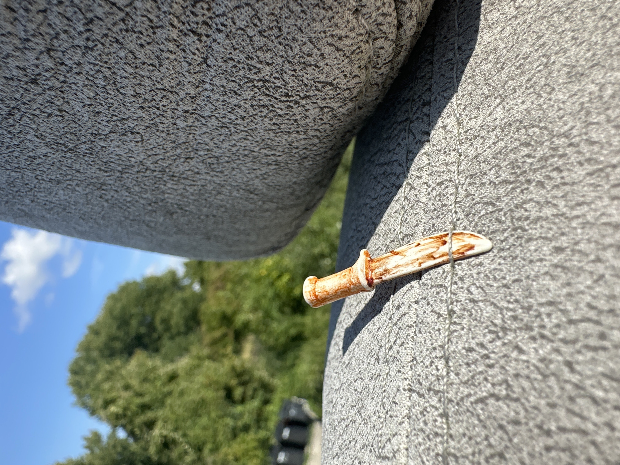 The image shows a miniature, ornamental dagger lying on a textured, grey surface, possibly a stone bench or monument. The dagger is made of a light-colored material, with brown staining that resembles blood. The background features lush green trees and a bright blue sky with a few white clouds, suggesting an outdoor setting. In the distance, some dark, cylindrical objects are visible, possibly bins or planters. The scene appears to be set in a park or a public space. This location is identified as being in Lelystad, Netherlands. The perspective is low, close to the ground, with the dagger as the primary subject.