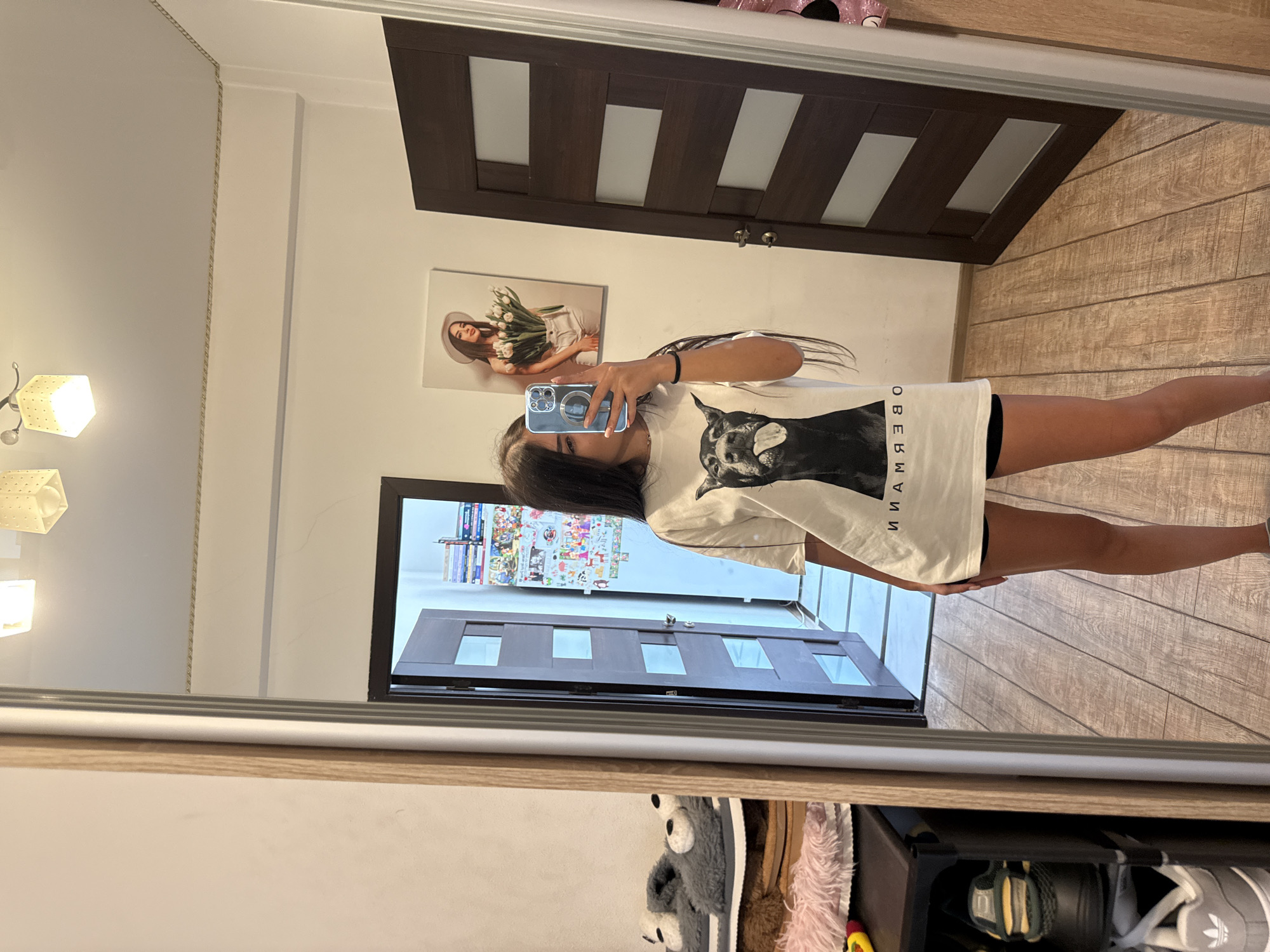A young woman with long dark hair is taking a mirror selfie in what appears to be a hallway or bedroom in a residential apartment. She is wearing a white t-shirt with a large black and white graphic of a dog's head and the word "OBERMANN" printed in black text, along with black shorts. She is holding a smartphone in her right hand to take the photo.
The background includes a large mirror, a closed dark brown wooden door with horizontal glass panels, and a wall with a framed picture of a woman holding a bouquet of flowers. To the left of the woman, in the reflection of the mirror, are shelves with various items, including plush slippers, fuzzy slippers, and shoes. The floor is made of light-colored wooden planks. The overall impression is a casual, indoor setting. The specific location in Chișinău, Moldova cannot be determined from the image.