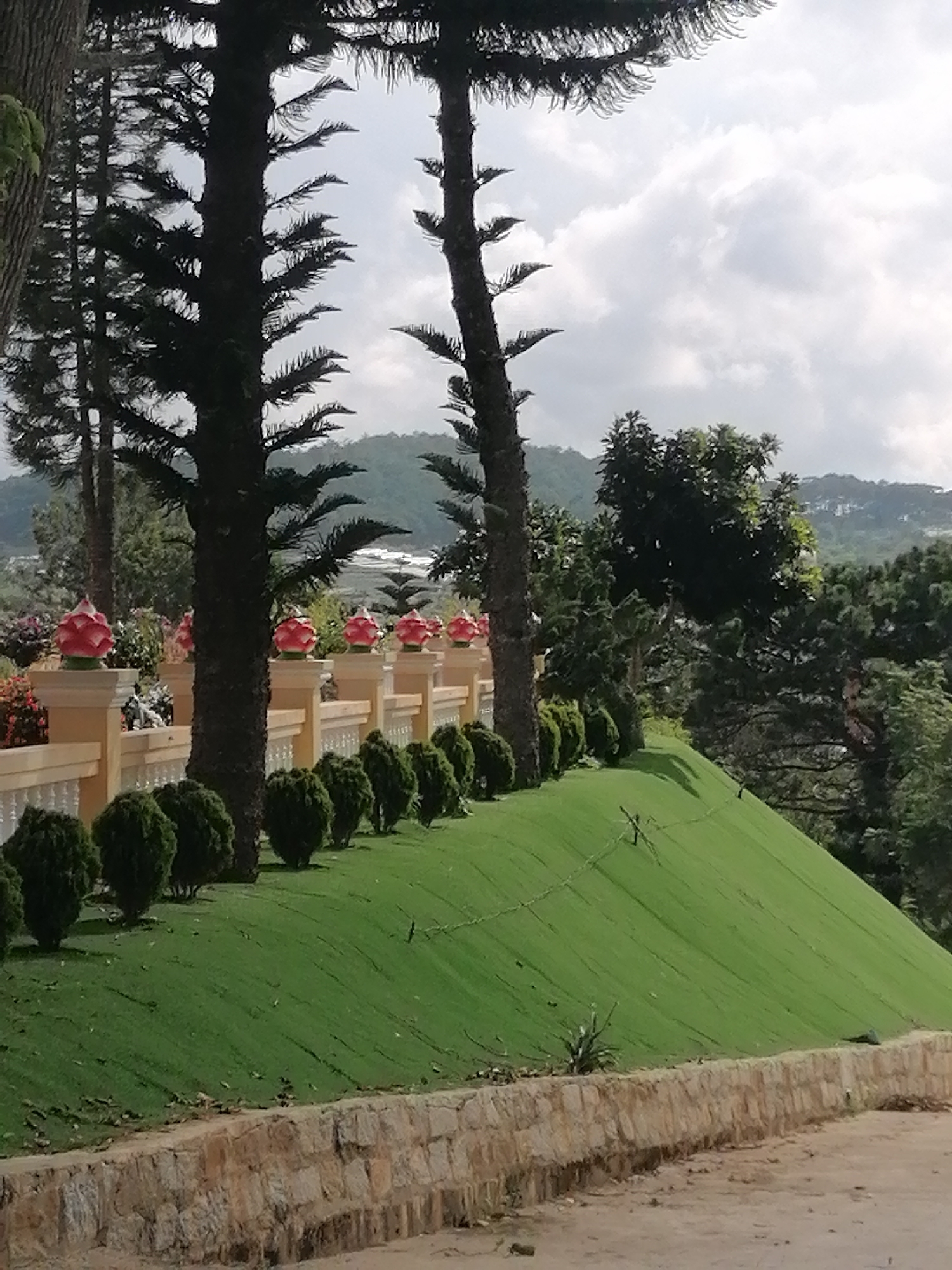 The image shows a landscaped area with a man-made hill covered in bright green artificial turf. A low stone wall borders the foreground, and a row of evenly spaced, dark green, rounded bushes runs along the top of the artificial turf hill. Behind the bushes is a pale yellow ornamental fence topped with red and white lotus-shaped finials. Tall, dark evergreen trees are prominent in the foreground and midground, with their branches reaching towards a cloudy sky. In the background, rolling hills covered in trees and vegetation are visible. The scene appears to be in an outdoor park or garden setting within Lộc Quý, Vietnam, with a focus on decorative landscaping and natural elements. No people are visible in the image.