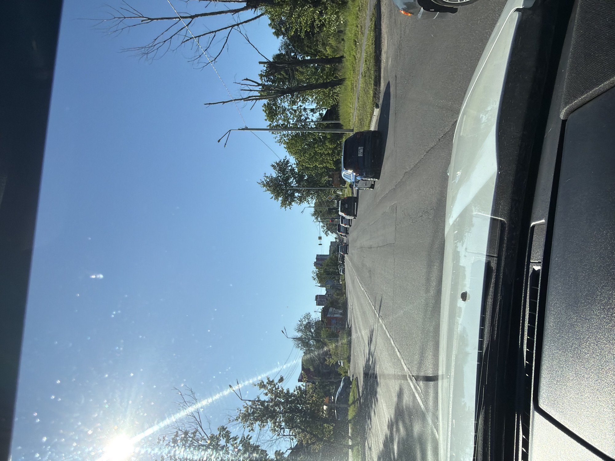 The image depicts a view from inside a vehicle, looking forward through the windshield onto a sunny street in Khabarovsk, Russia. The sky is a clear, bright blue. In the distance, several multi-story buildings are visible, suggesting an urban or suburban setting. The road is paved with asphalt and has lane markings. Several cars are driving on the road, some of which are dark-colored sedans. Trees and utility poles line the sides of the road. The sun is bright and shining from the left, casting a glare on the windshield. The interior of the vehicle is partially visible in the foreground, including the dashboard and part of the hood.