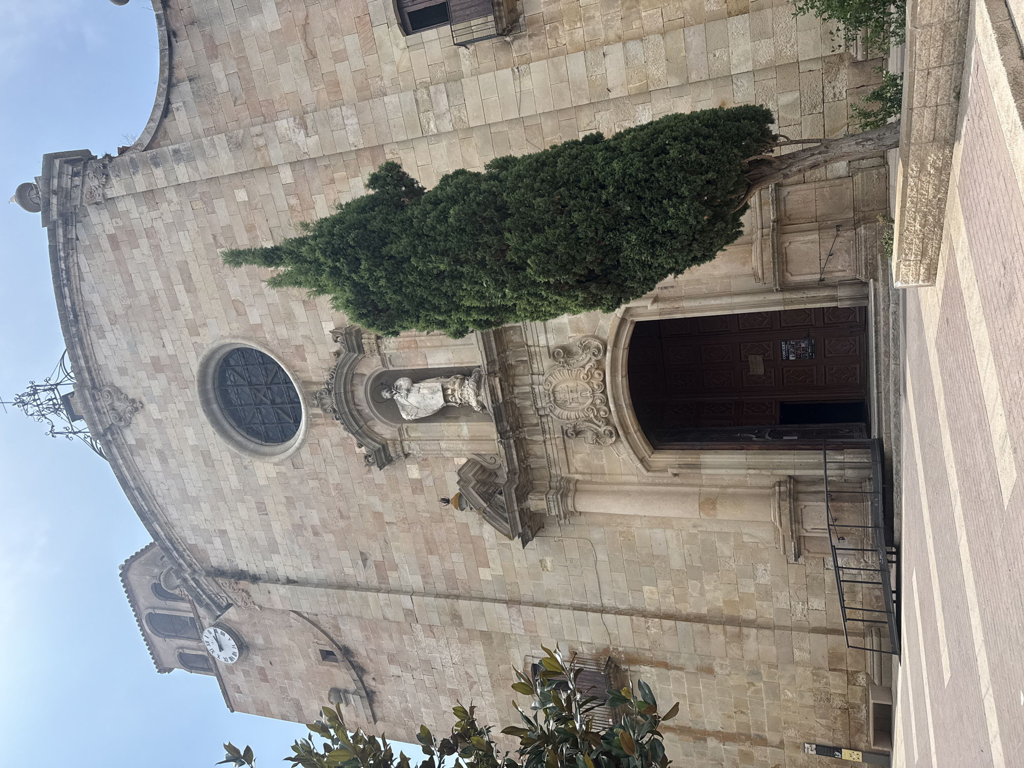 This is a full, eye-level shot of the façade of the Church of Our Lady of the Sea in Tossa De Mar, Spain. The church is constructed of tan-colored stone blocks. The façade features a large, dark wooden double door with intricate carvings, accessed by a ramp. Above the door is a decorative stone arch and a niche containing a statue of a robed figure. To the left of the door, a clock face with Roman numerals is visible on a square tower. A large, slender, dark green evergreen tree stands prominently to the right of the entrance, partially obscuring the church's central architectural features. The sky is a pale blue, indicating a clear day. In the foreground, a patterned brick pavement leads up to the church entrance. A portion of a green tree with broad leaves is visible in the upper left corner, and a balcony railing can be seen on the right side of the church.