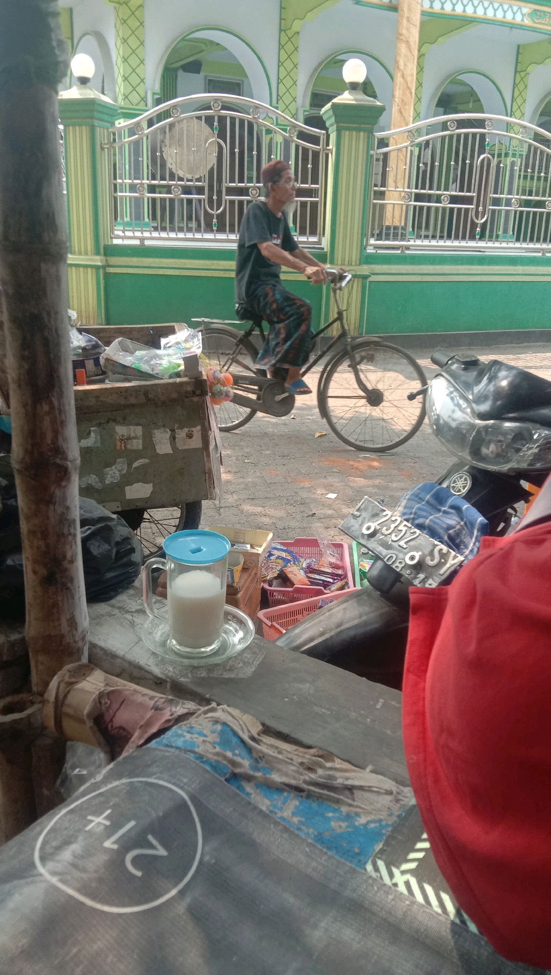In Ngempit, Indonesia, a man with grey hair and a beard is riding a bicycle. He is wearing a dark t-shirt and patterned pants. The bicycle has a rear rack with colorful decorations. The man is riding past a green and white building with arched windows and decorative metal fencing. In the foreground, there is a glass of milk with a blue lid on a saucer. Next to it is a cart with various items, including plastic bags and what appears to be merchandise. A portion of a red garment and a motorcycle are also visible in the foreground. The scene appears to be an outdoor market or roadside setting.