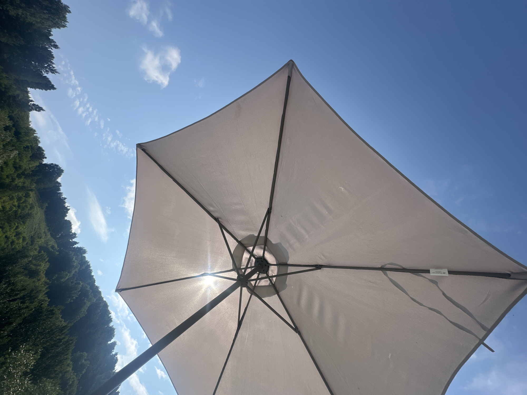 The image shows a close-up, upward-facing view of a light-colored rectangular umbrella. The umbrella's fabric is taut, with visible metal ribs supporting it. Sunlight is shining through the fabric in the center, creating a lens flare effect. The sky is a clear, bright blue with a few scattered white clouds. In the background, a tree-covered hillside is visible, suggesting a natural or semi-rural environment. There are no people or animals in the frame. The scene appears to be outdoors on a sunny day. The location is stated to be Danilovka (Altai), Russia.