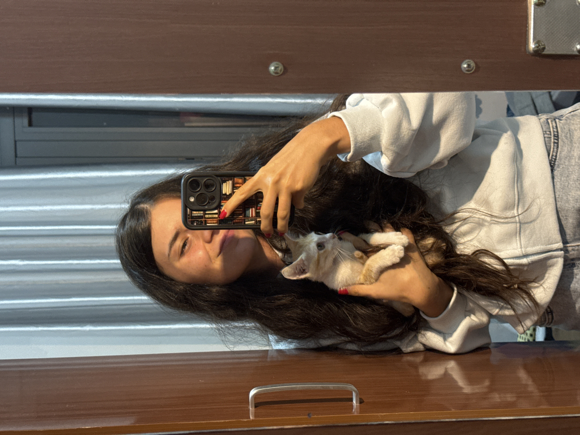 A young woman with long, dark hair is reflected in a mirror, holding a small, light-colored kitten. She is wearing a light blue hooded sweatshirt and blue jeans. The kitten is held in her left arm and appears to be light orange and white with light blue eyes. The woman is taking a selfie with her phone, which has a case decorated with a bookshelf pattern. She has a slight smile on her face and her fingernails are painted red. The background shows vertical blinds and a wooden closet door with a handle and a circular metal fixture. The setting appears to be indoors. There is no indication of the city of Mũi Né, Vietnam in this image.