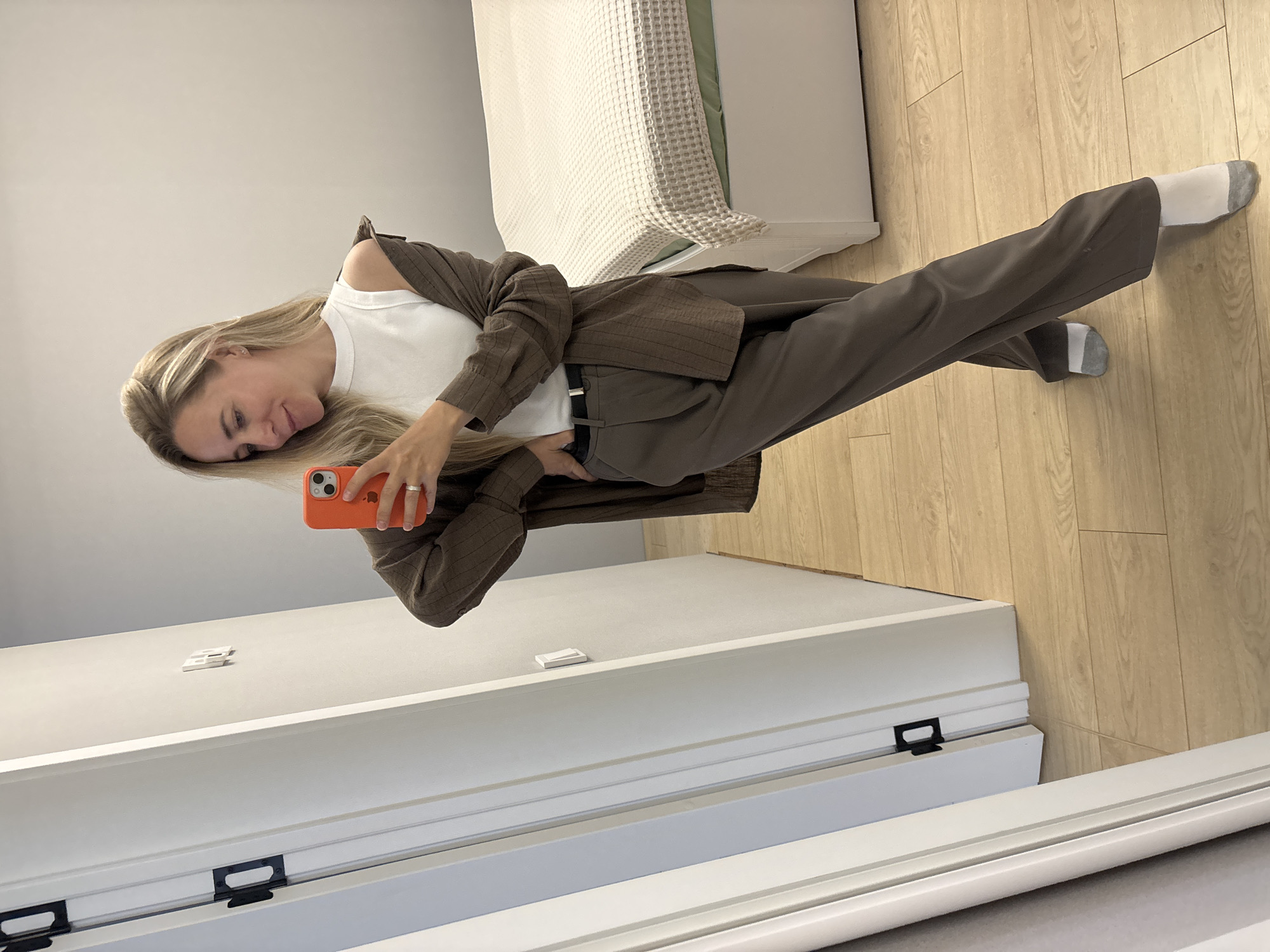 A woman is standing in a room and taking a selfie in the mirror. She is wearing a white sleeveless top, brown patterned blazer draped over her shoulders, and brown trousers with a belt. She has blonde hair and is wearing white socks with grey accents. The room has light-colored walls, a wooden floor, and a bed with a white textured comforter. A white door is visible on the left side of the image. The scene appears to be an indoor residential setting, possibly a bedroom or dressing area, located in Chaikovsky, Russia.