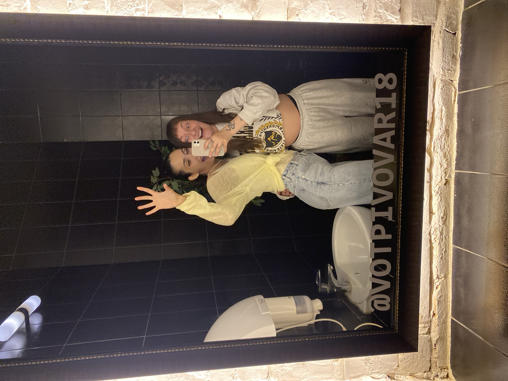 Two women are reflected in a large, ornate mirror in a dimly lit restroom. The woman on the left is wearing a yellow sheer long-sleeved shirt tied at the waist and light-wash denim jeans, with her left arm raised and hand waving. The woman on the right is wearing a grey cropped sweatshirt and grey sweatpants, holding a white smartphone to take a selfie of herself and her friend. Both women have expressive faces, with the woman on the right making a surprised face with wide eyes and an open mouth, and the woman on the left smiling widely with her mouth open. The restroom has black tiled walls and a white sink with a chrome faucet. A white paper towel dispenser is mounted on the wall to the left of the sink. The bottom of the mirror has the text "@VOTPIVOVAR18" etched into it. The overall environment appears to be a bar or club in Votkinsk, Russia. The lighting is dim, with a bright overhead light creating glare on the mirror and a warm light source illuminating the brick wall to the right of the mirror.