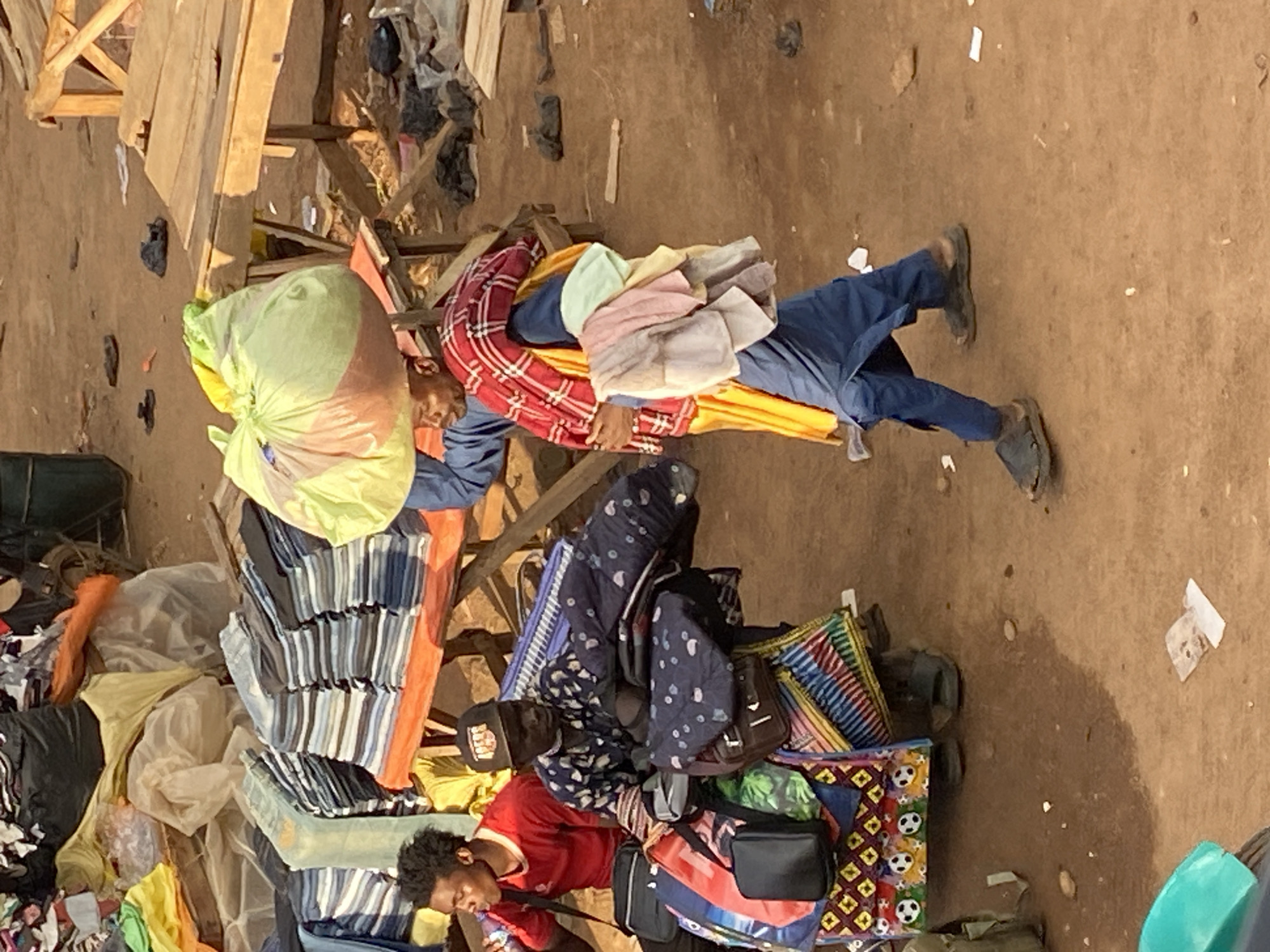 This image depicts a segment of an outdoor market in Bukuru, Nigeria. The ground consists of reddish-brown, uneven dirt, scattered with debris including small pieces of paper, plastic, and discarded dark sandals.
Three adult males are visible within the frame. On the left, a young man with dark, curly hair, wearing a red short-sleeved shirt and a black sling bag, is seated and appears to be drinking from a clear plastic bottle. In the center, an older man, wearing a dark patterned long-sleeved shirt and a black baseball cap with "49ERS" printed on it, is seated behind a multi-layered cart. This cart is laden with various bags and folded textiles, including a blue bag with white polka dots and a lower section with a yellow background and black soccer ball patterns.
On the right, a third man is walking from left to right. He carries a large, light green/yellowish bag on his head. He is dressed in a blue outer garment over a yellow one, with a red and white plaid fabric draped over his left shoulder and a stack of folded cloths (pink, light green, yellow, grey) balanced on his left arm. He is wearing dark sandals on his feet.
In the background, on the left, an orange-covered market stall table displays neatly folded stacks of clothing, including jeans and various colored shirts. Further back, more jumbled textiles are piled. On the right, a rough wooden table structure, likely part of another stall, is present. The scene is illuminated by strong, directional sunlight, indicating an outdoor daytime setting.
