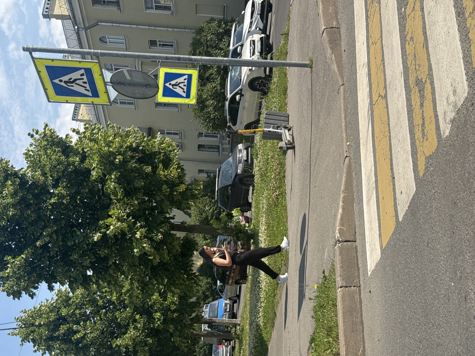 A woman walks on a sidewalk next to a street with parked cars and trees. She is wearing black pants and a black sleeveless top. She is looking at her phone, which she is holding in her left hand. A scooter is parked on the sidewalk near a pedestrian crossing sign. The pedestrian crossing sign is yellow and blue and shows a silhouette of a person walking. The street is paved with asphalt and has white and yellow stripes for a pedestrian crossing. The buildings in the background are light-colored and have arched windows. The sky is blue with some clouds. The scene appears to be in Metallostroy, Russia, a city known for its industrial and residential areas.