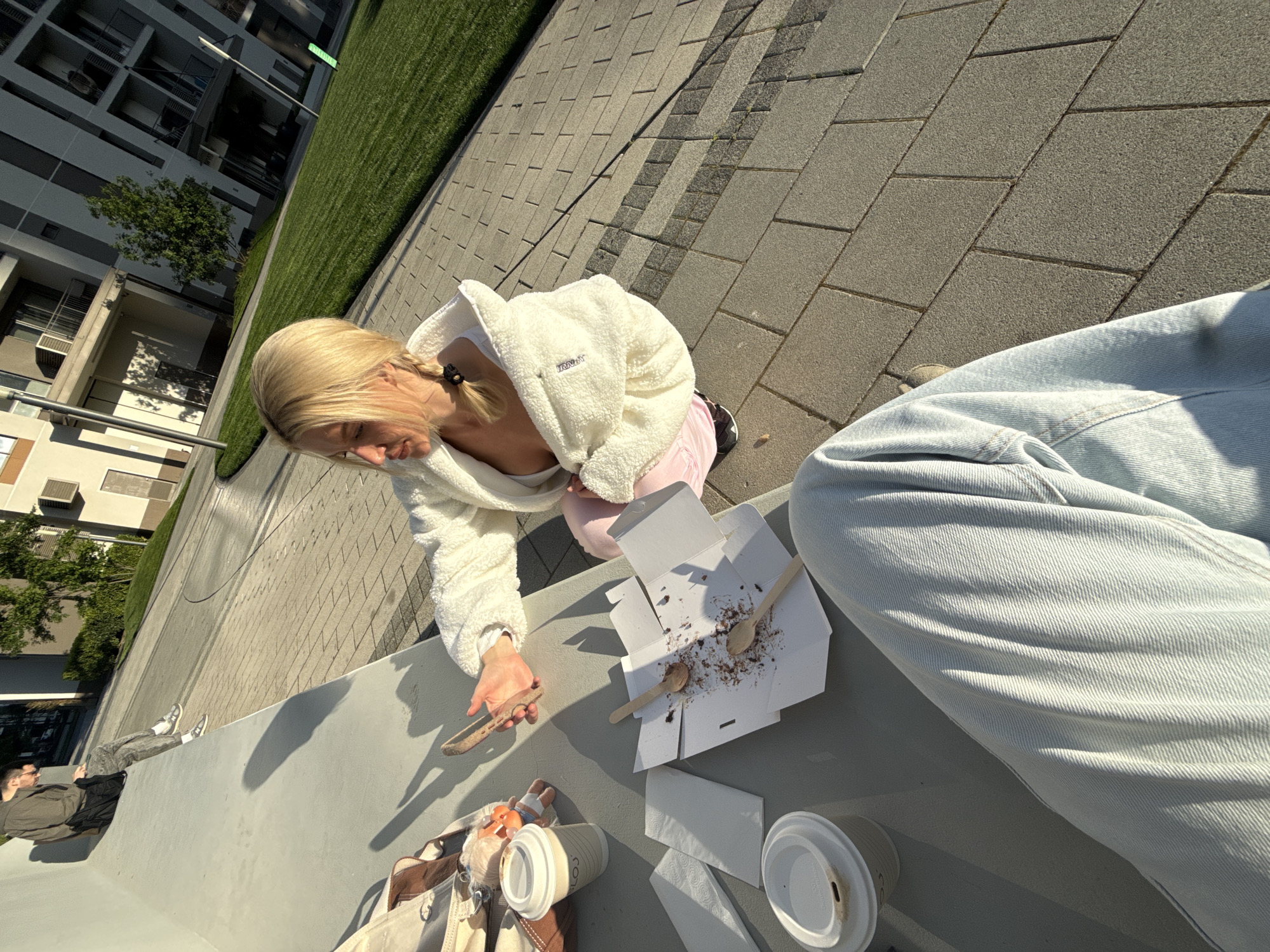 The image captures a casual, sunlit outdoor scene in an urban environment, identified as Belgrade, Serbia.
In the foreground, a blonde woman, wearing a white fluffy jacket over pink pants, is leaning over a concrete ledge. Her hair is tied back in a ponytail with a small braid. She holds a dull-edged knife-like utensil, appearing to scrape the last bits from an open white cardboard dessert box. The box contains chocolate crumbs and two wooden spoons. Nearby on the ledge are two paper coffee cups with lids, white napkins, a brown and cream-colored tote bag, and a small doll or toy. The leg of another person, wearing light blue jeans, is visible in the bottom right corner, suggesting a shared activity or a first-person perspective.
The background features modern, multi-story buildings with balconies, a strip of manicured green grass, and paved areas with large rectangular tiles. Further back, a man wearing dark clothing and sunglasses is seen sitting on a similar concrete ledge, facing away. A tree and a street light are also visible.
The strong, distinct shadows indicate a bright, sunny day, likely morning or late afternoon. The overall atmosphere is relaxed and informal, suggesting friends enjoying a snack or coffee break outdoors. Faint text, possibly "TRNHT," is visible on the woman's white jacket.