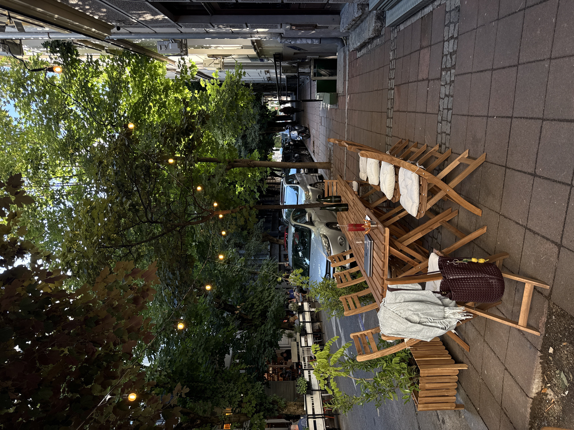 The image shows a street scene in Belgrade, Serbia. In the foreground, a wooden table with chairs is set up on a paved sidewalk, suggesting an outdoor seating area for a cafe or restaurant. The table is set with a glass of red liquid, a laptop, and several bottles. A grey throw blanket and a dark woven bag hang from one of the chairs.
Behind the table, a street runs alongside the sidewalk, with several cars parked on the street. A silver sedan is prominently visible. Trees line the street, providing shade, and strings of lights are strung between them, indicating a nighttime or evening ambiance.
Further down the sidewalk, more tables and chairs are visible, along with people seated at them, enjoying the outdoor space. Buildings line the opposite side of the street. The overall impression is of a lively urban environment with an emphasis on outdoor dining and social gathering.