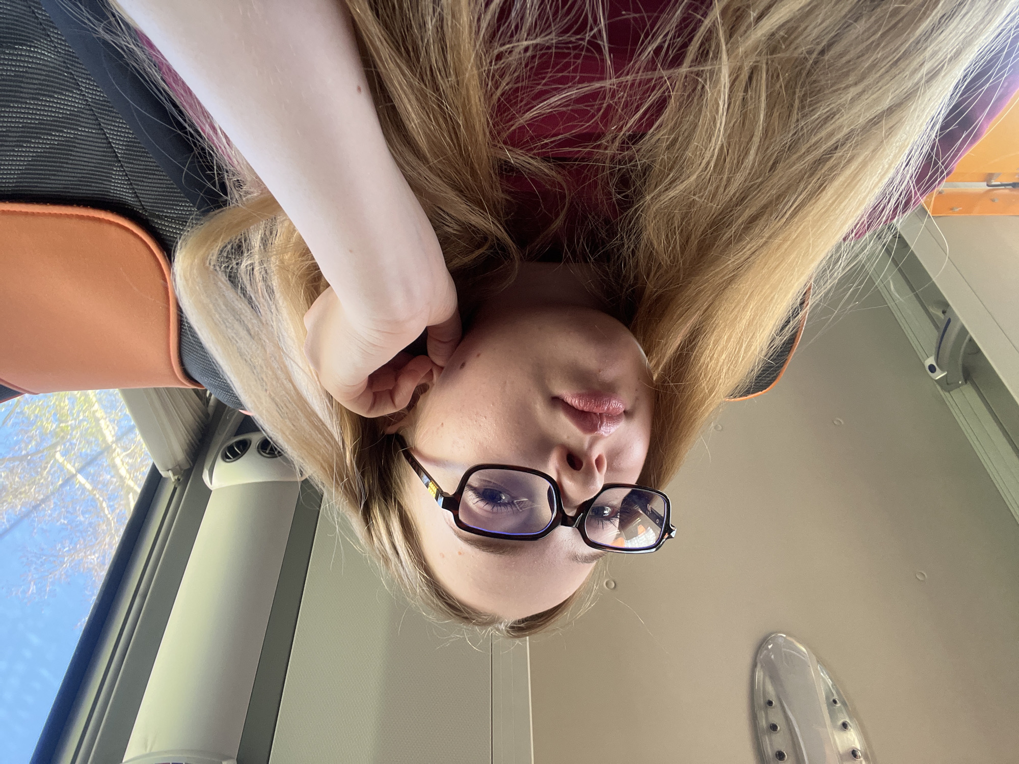 A blonde-haired individual wearing glasses is presented in an inverted portrait orientation. The person is positioned inside a vehicle, potentially a bus, as suggested by the presence of a window and overhead fixtures. The background is a ceiling with several small indentations. The individual is holding their hand up to their ear. The location is unidentifiable as Chaikovsky, Russia.