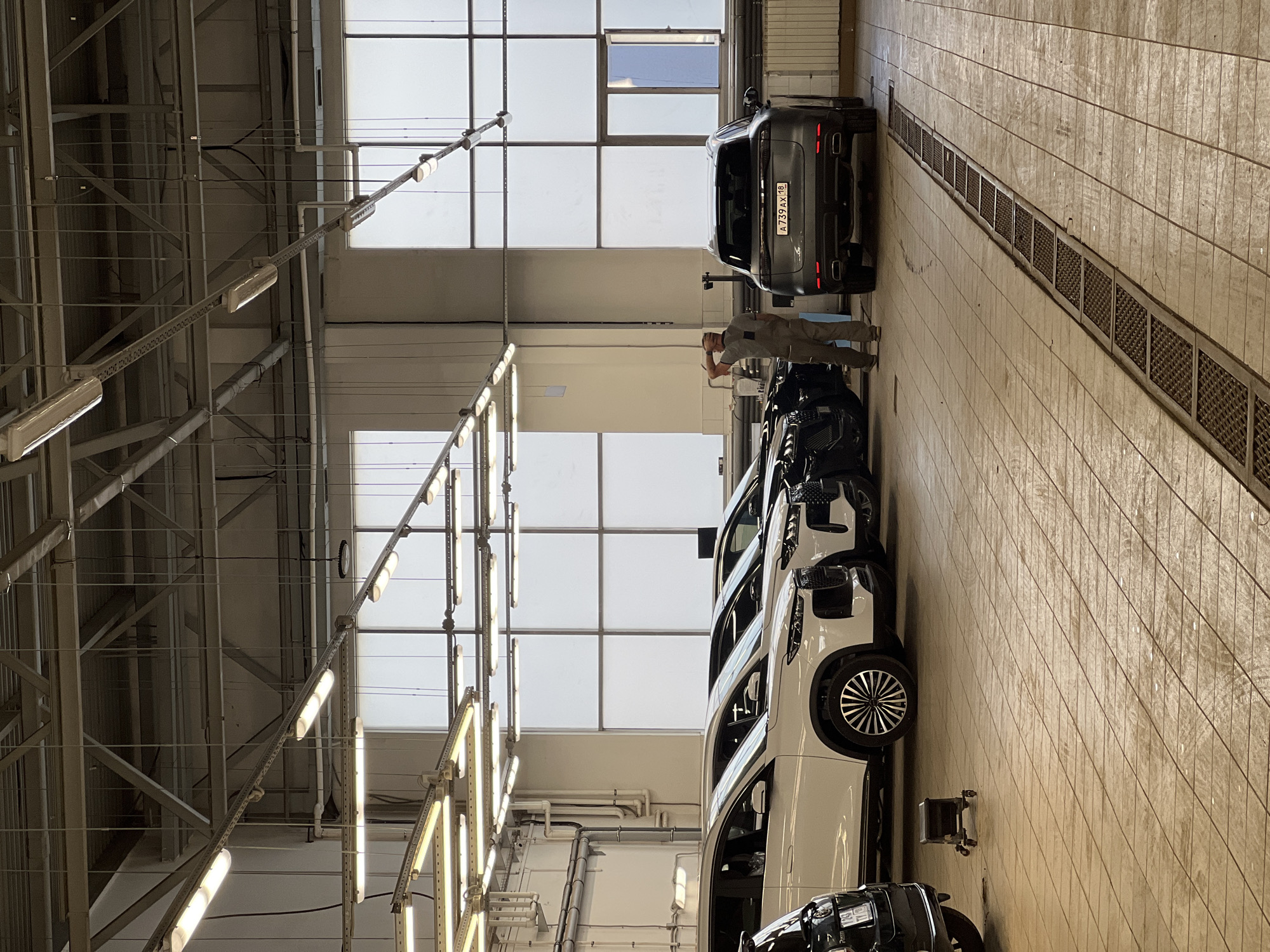 This image depicts the interior of a vehicle service or display area in Izhevsk, Russia. The space is illuminated by an extensive overhead grid of linear fluorescent light fixtures suspended from a metal truss ceiling structure, supplemented by ambient light diffusing through large, multi-paned windows that line the back wall. The floor is covered with light-colored square tiles, showing visible wear and minor debris, and features a long, recessed metal drainage grate running through the central foreground.
Multiple vehicles are present. On the left, a row of four modern SUVs is parked, facing left. The three foreground vehicles are white, exhibiting contemporary design features including large grilles and multi-spoke wheels. A fourth black SUV is partially visible behind the last white vehicle. On the right, a dark grey Porsche Cayenne SUV is parked with its rear facing the viewer. Its Russian license plate, "A 739 AX 18", is distinctly visible, confirming the Udmurt Republic where Izhevsk is the capital.
A single male individual stands near the rear of the grey Porsche Cayenne, his back mostly to the viewer. He is dressed in a light grey work shirt and lighter-colored work trousers. His right hand is placed on the back of his head, indicating a contemplative or observing posture. A tall, dark, slender object resembling a stand is positioned behind him, adjacent to the SUV. In the lower left foreground, a small, dark-colored wheeled creeper or stool rests on the floor. White pipes and conduits are visibly mounted along the left wall.